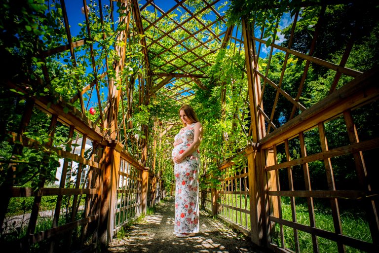 Pregnant woman standing on wooden pergola walkway covered in climbing greenery and sunlight.