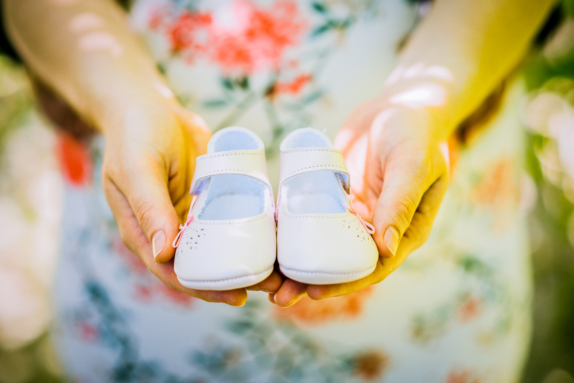 Close-up of hands holding tiny baby shoes against floral maternity dress in warm sunlight.