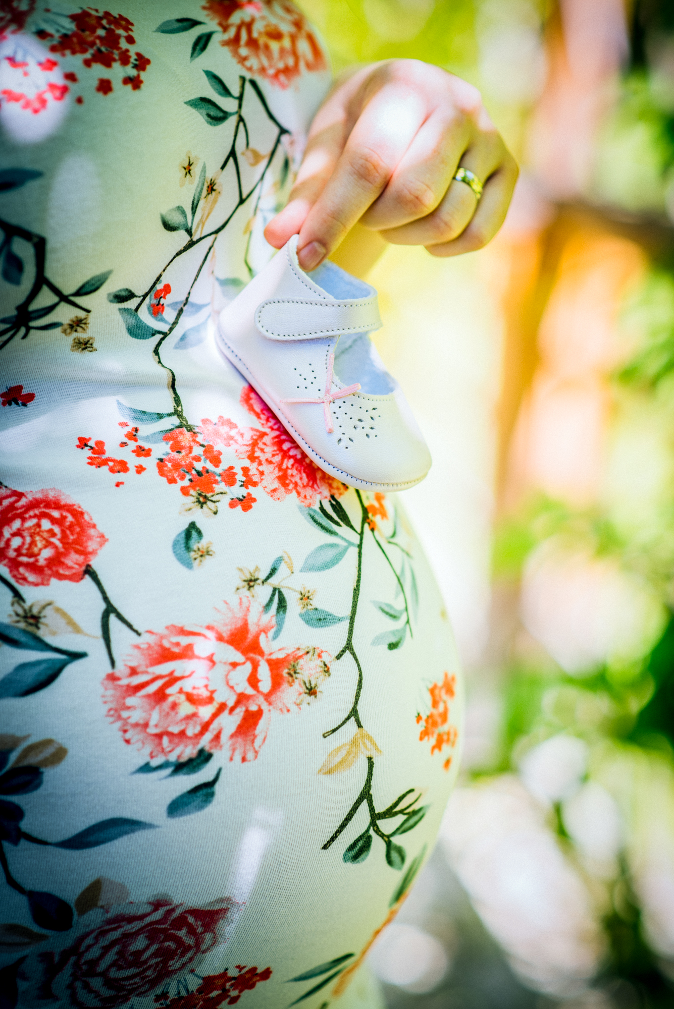 Detail of baby shoe held near pregnant belly, highlighting soft textures and floral pattern.