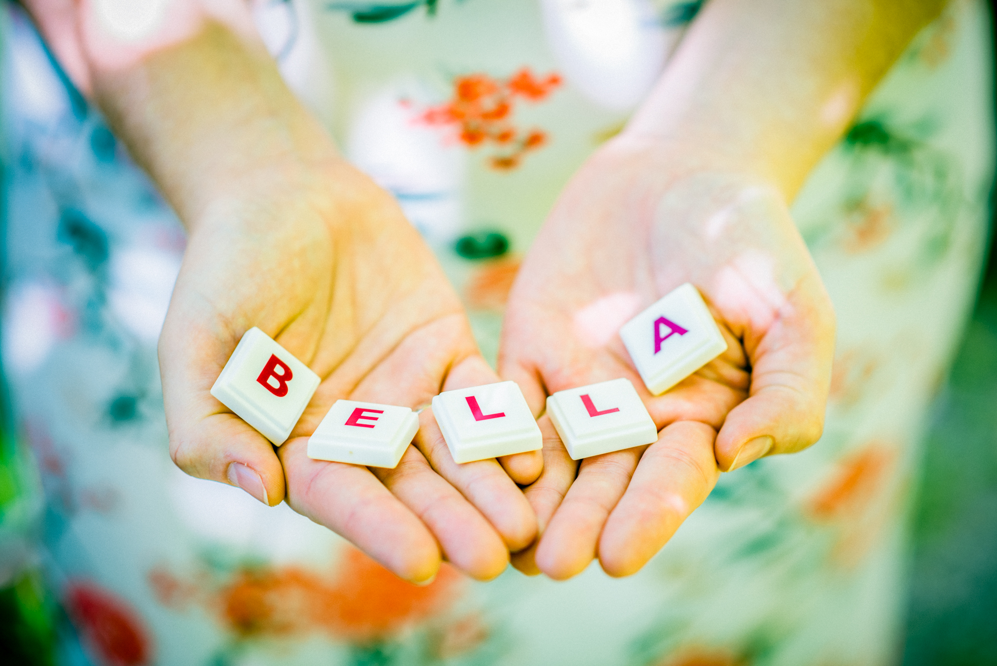 Hands holding letter blocks spelling “Bella” against floral dress during maternity session.