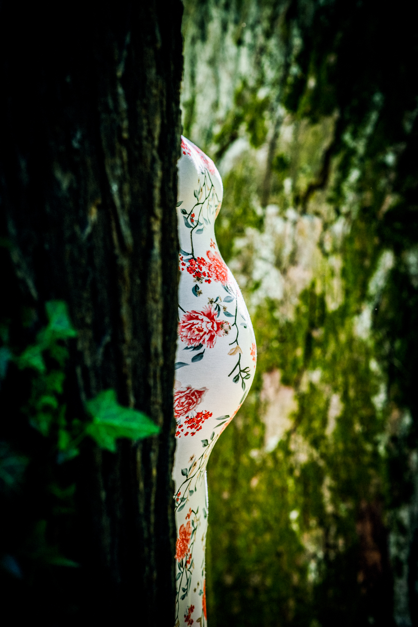 Close side view of pregnant belly in floral dress partially hidden behind tree trunk.