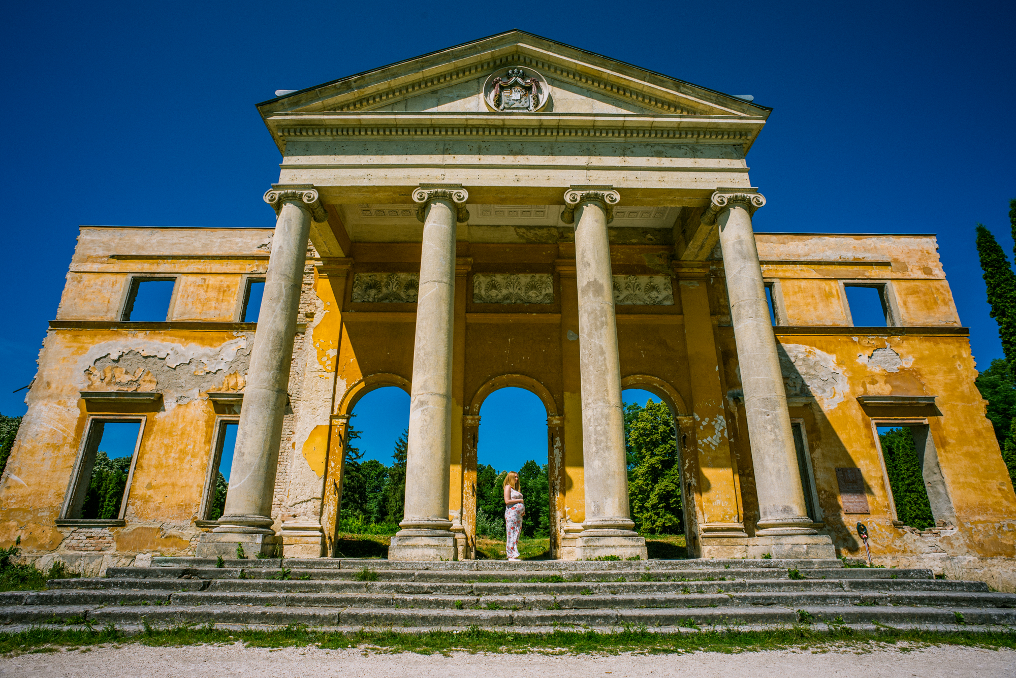 Pregnant woman framed by grand yellow ruin with columns at Vácrátót Botanical Garden.