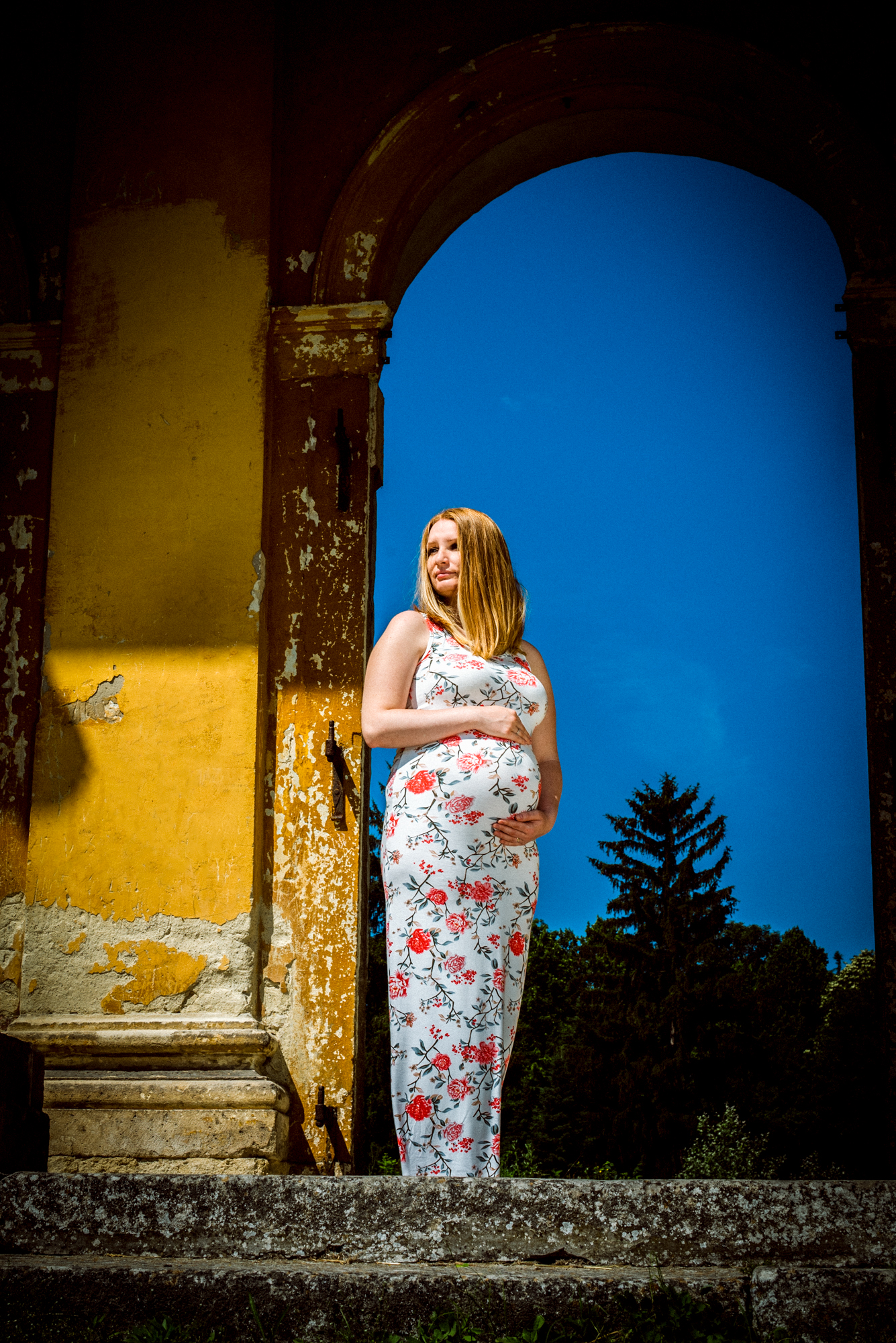 Expectant mother standing in arched doorway of historic ruin against deep blue sky.