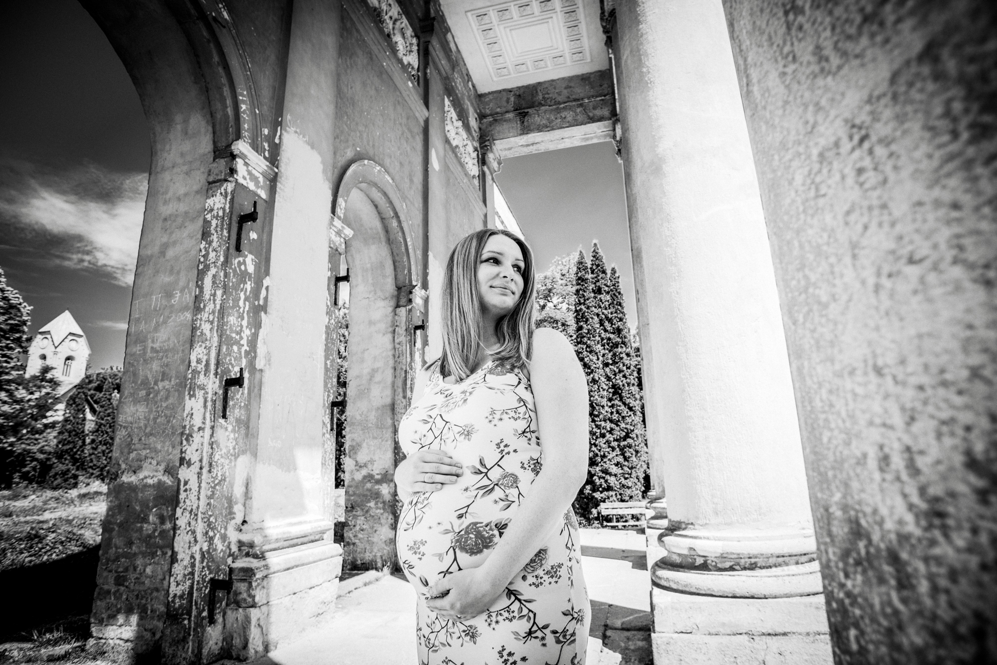 Pregnant woman standing among blooming garden plants with wooden trellis structure behind her.