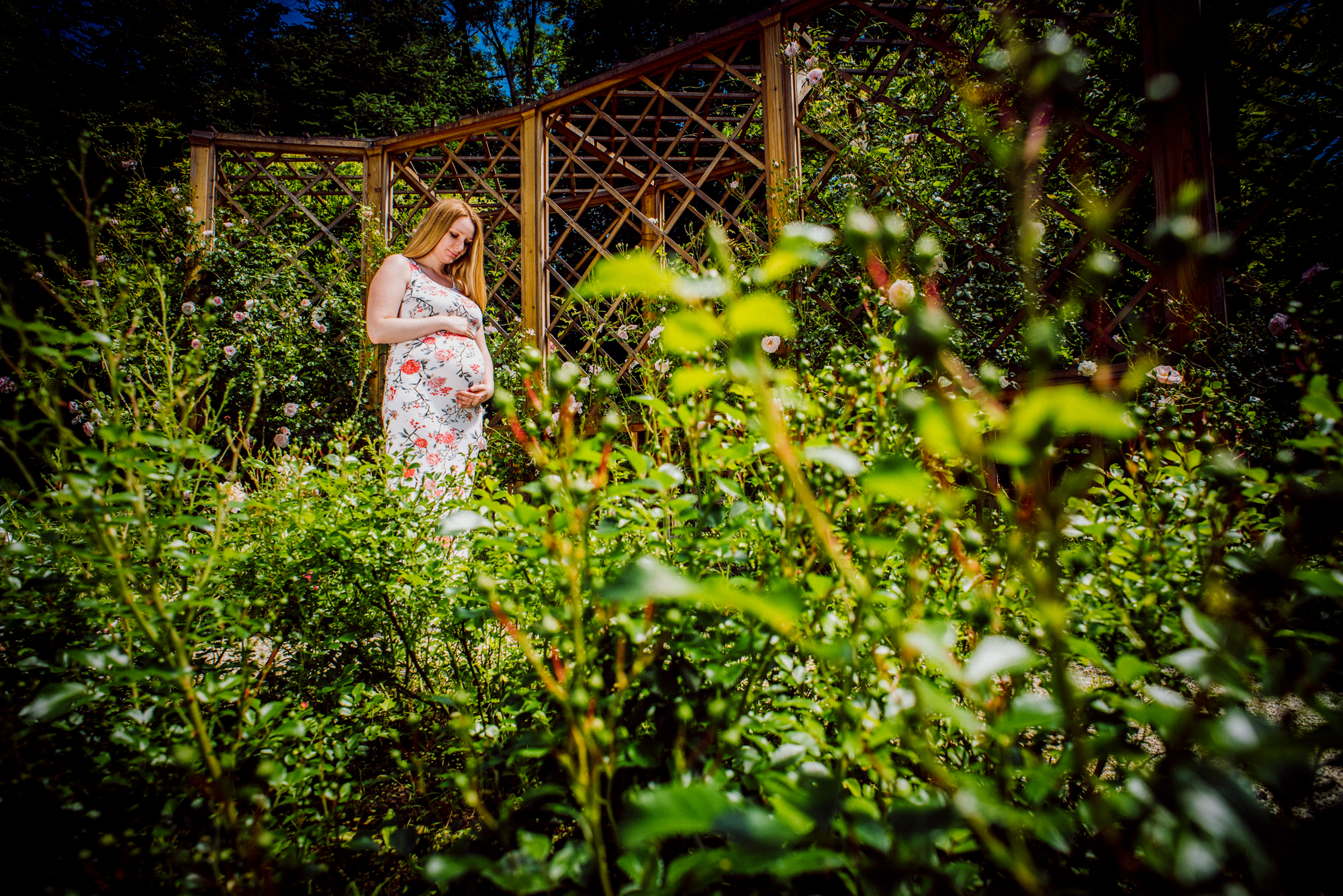 Expectant mother in floral dress surrounded by greenery and flowers in botanical garden setting.