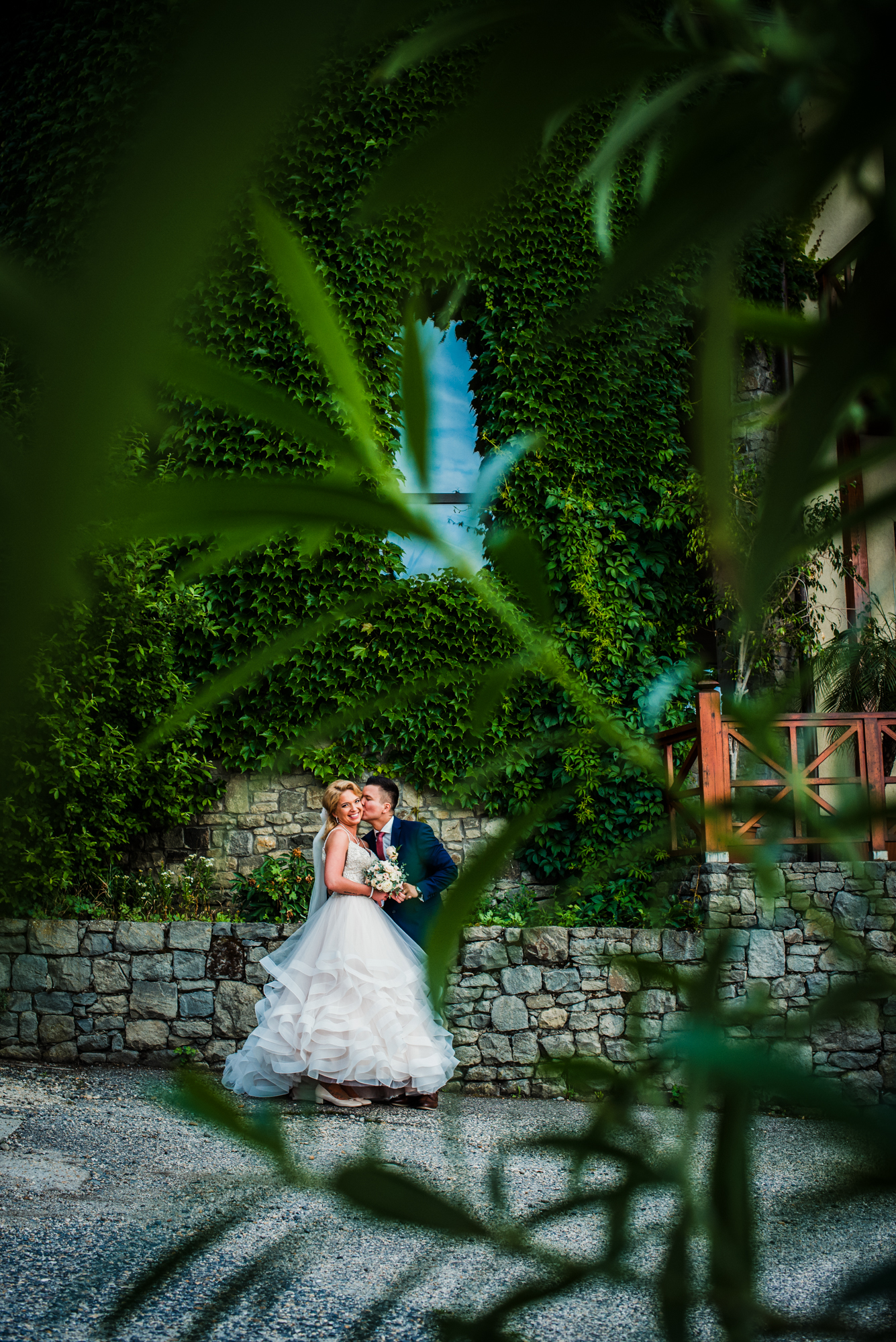 Couple standing by ivy-covered stone wall framed through leaves.