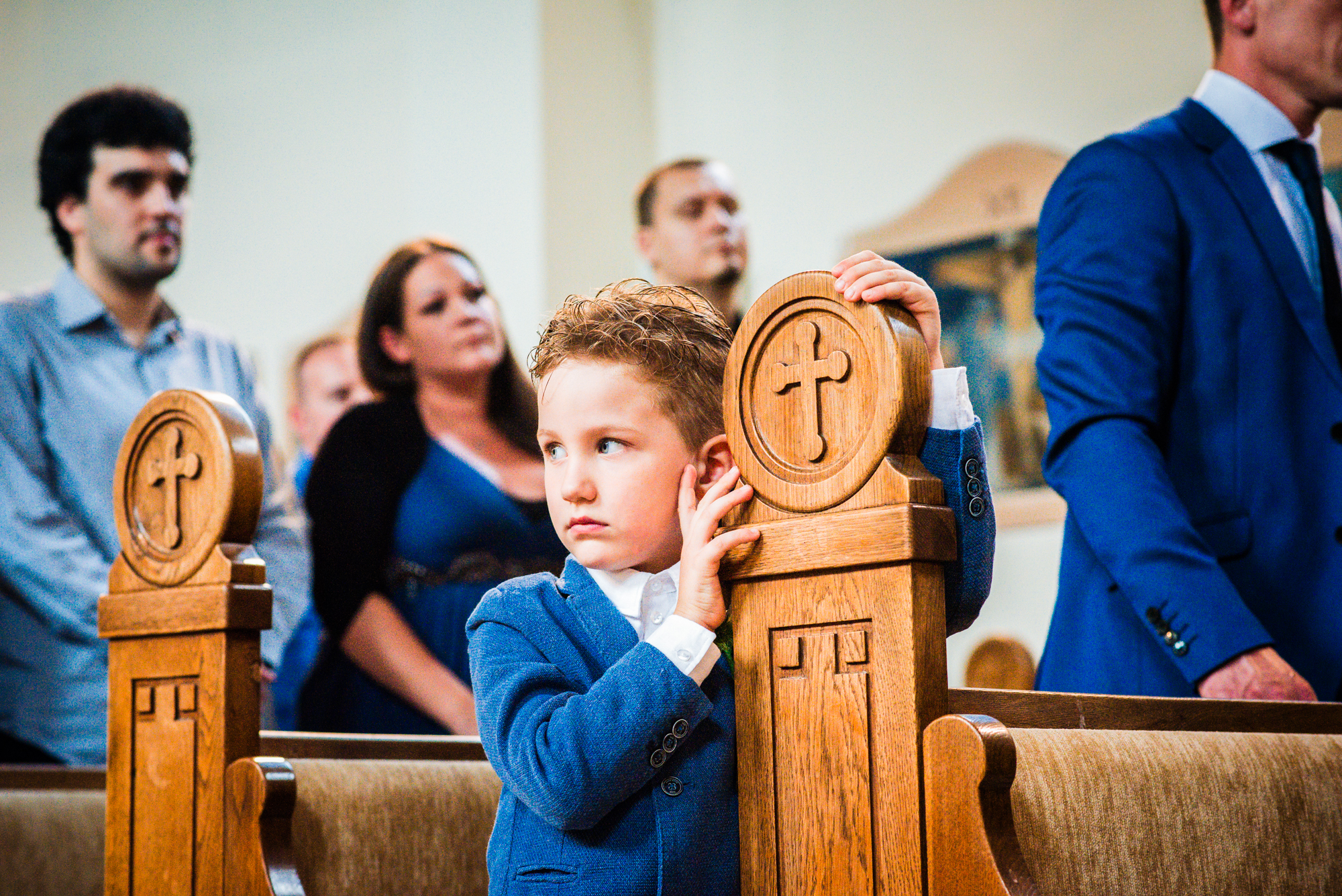 Boy in blue suit leaning on wooden pew, observing ceremony quietly with thoughtful expression.