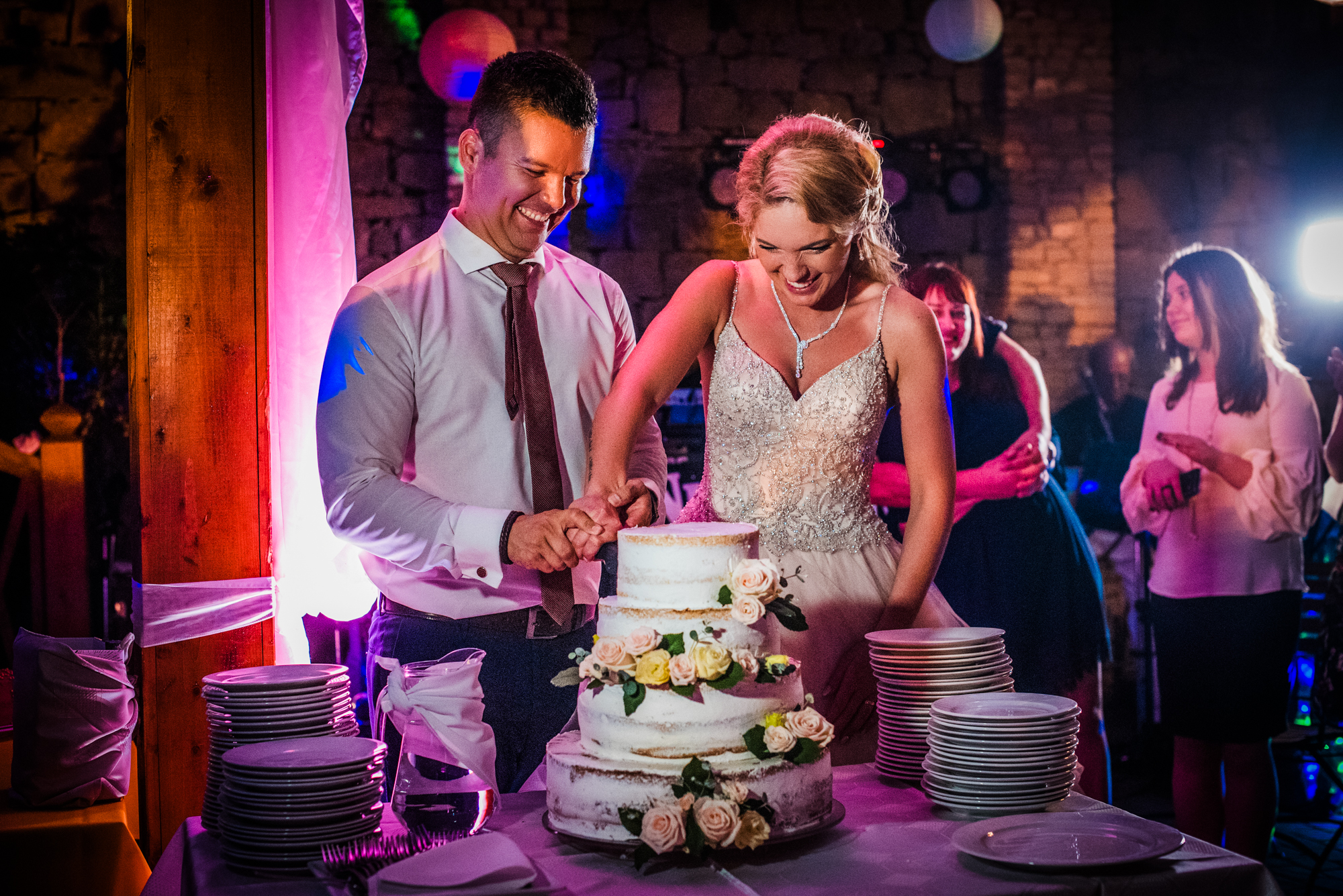 Couple cutting wedding cake together during reception with guests watching.