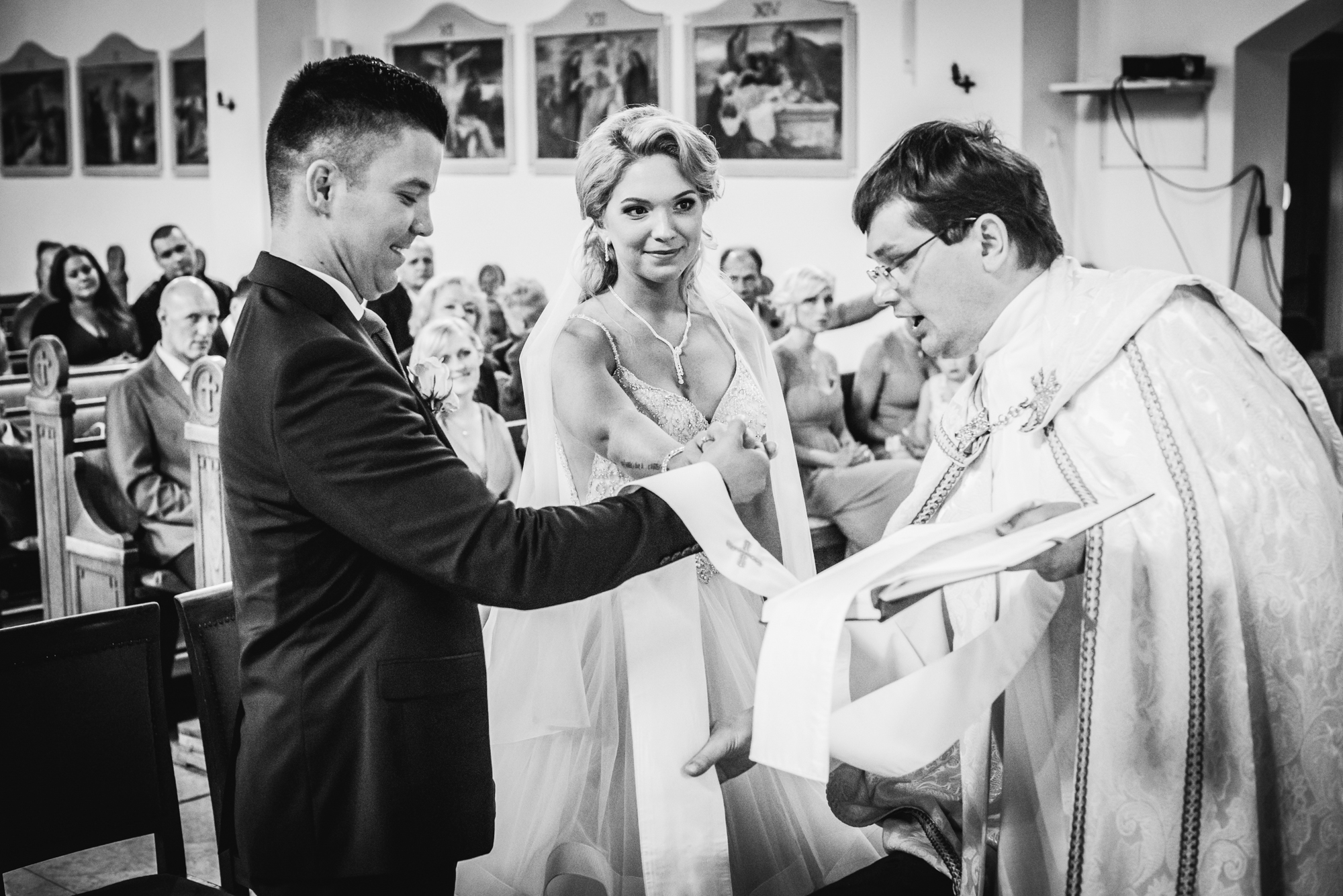 Couple exchanging vows with priest, hands joined as ceremony progresses in intimate church setting.