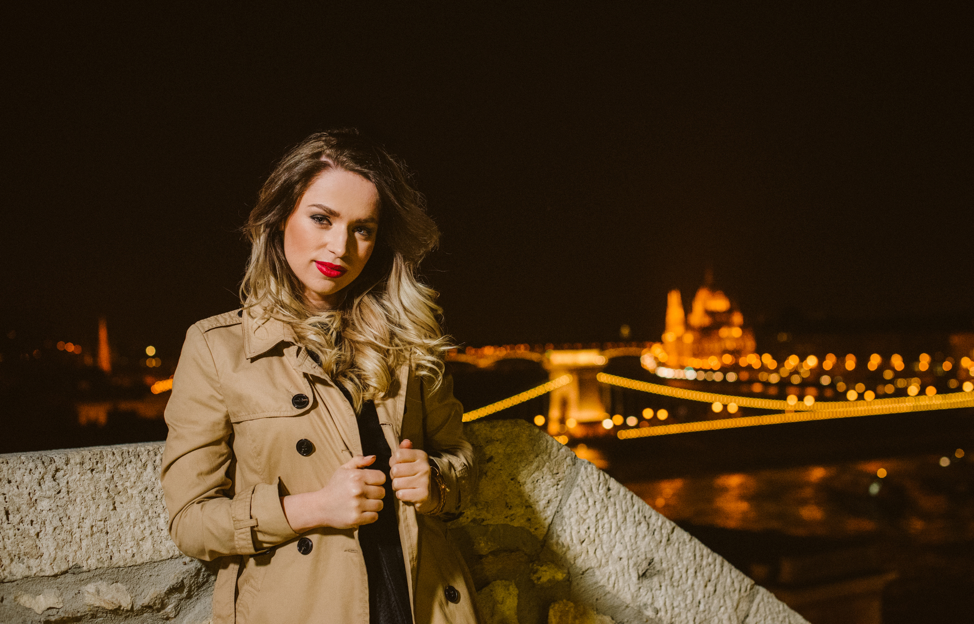Night portrait with trench coat and Budapest skyline lights behind.