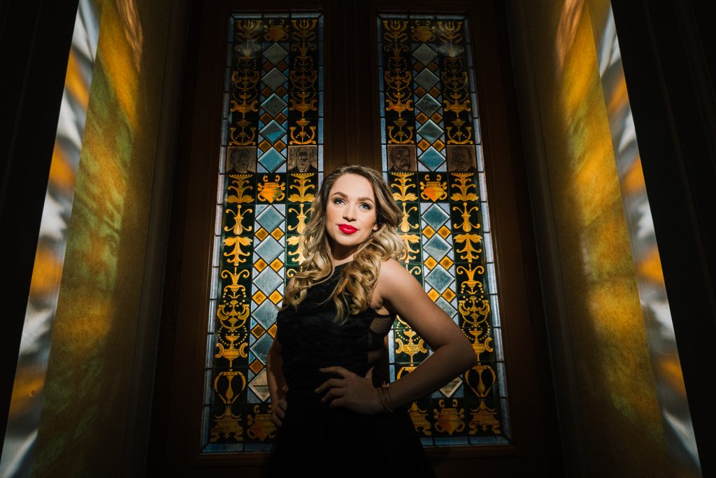 Woman standing before stained glass window with gold and green tones.
