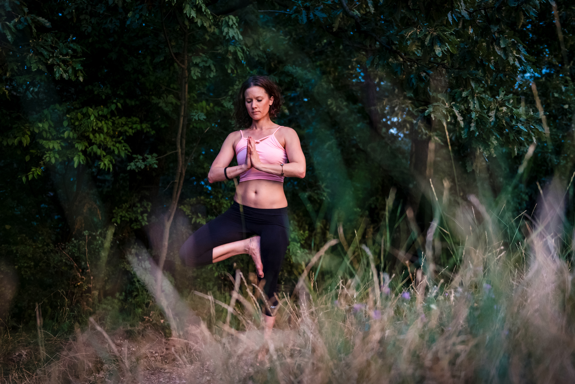Yoga teacher balancing outdoors behind soft blurred grass during an evening wellness photoshoot.