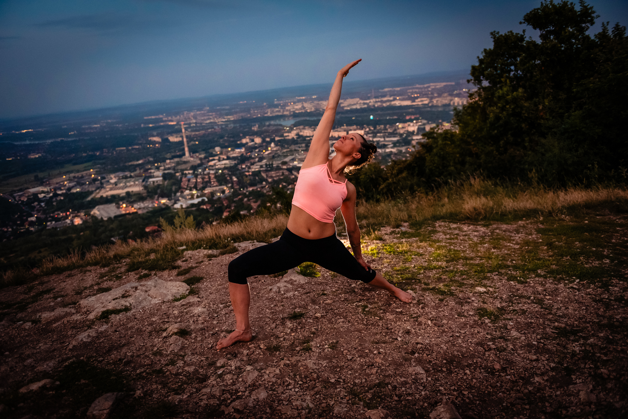Woman performing reverse warrior pose above Budapest city lights at blue hour on Three Border Mountain.