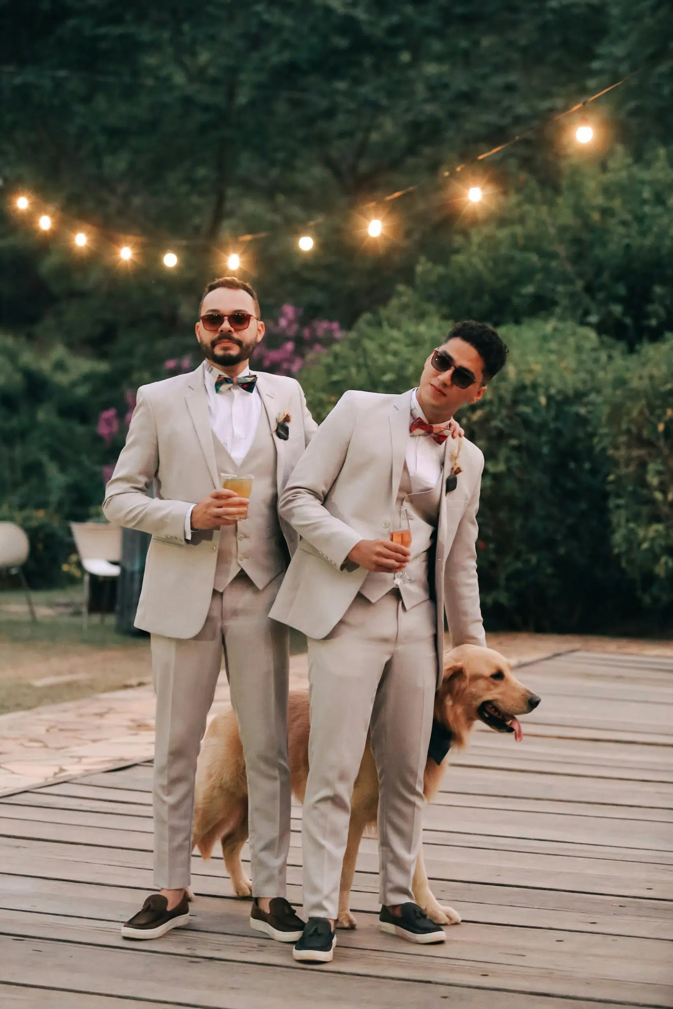 Two grooms in suits with their dog during outdoor wedding celebration under string lights