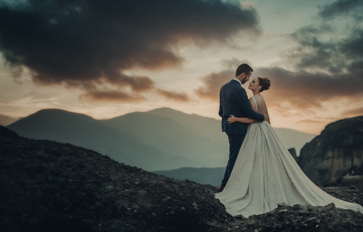 Bride and groom embracing on rocky peaks at sunset with dramatic mountain backdrop.