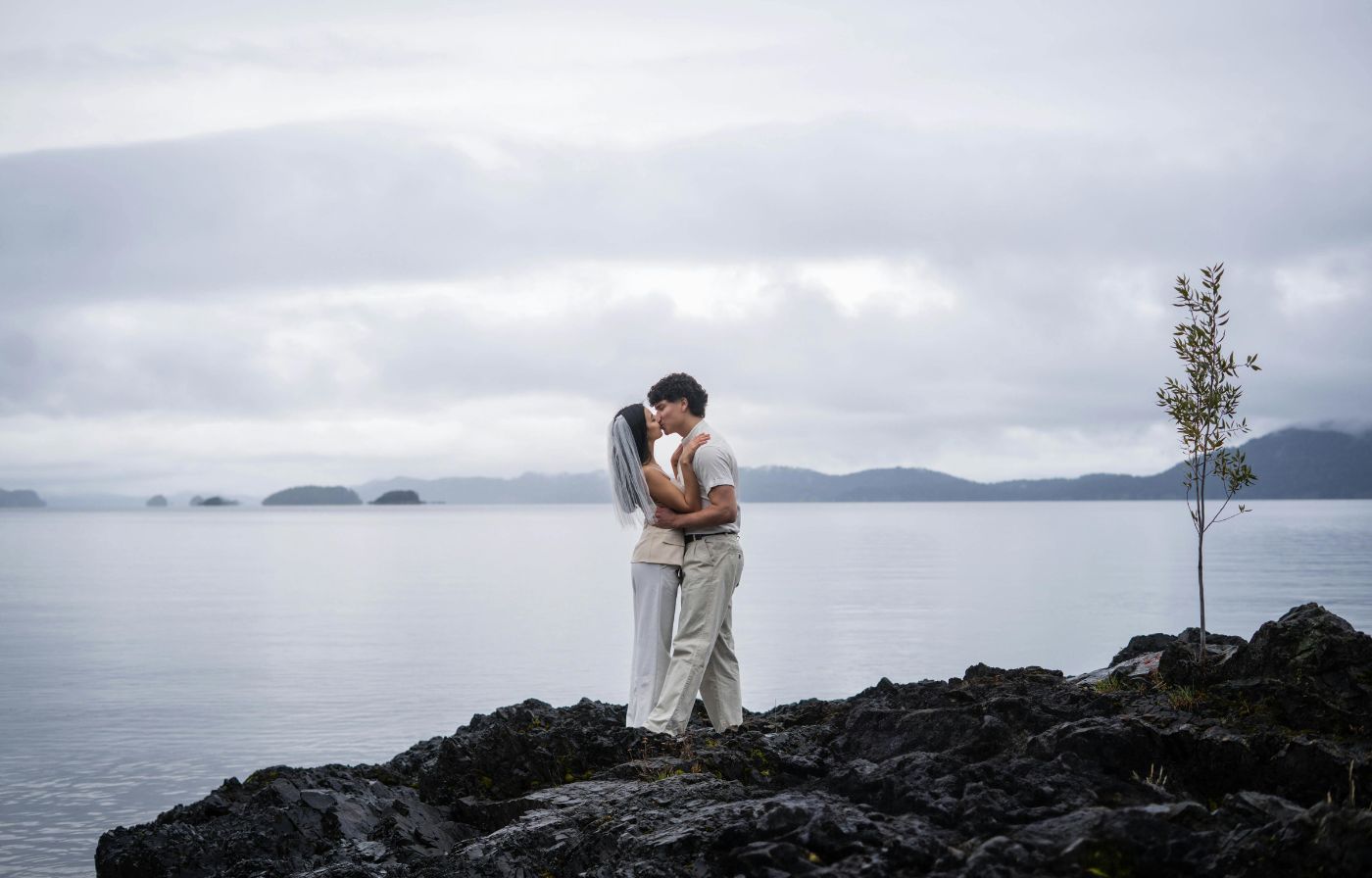 Bride and groom kissing on black rocks beside a misty lake.