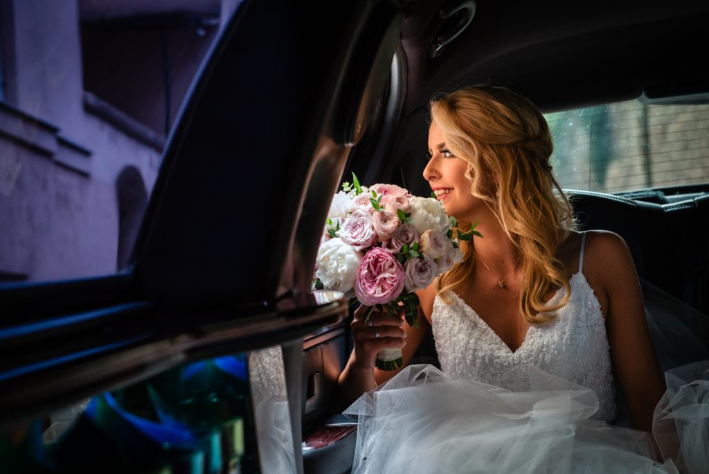 Bride sitting in a wedding car, smiling softly while holding a pastel bouquet before the ceremony.