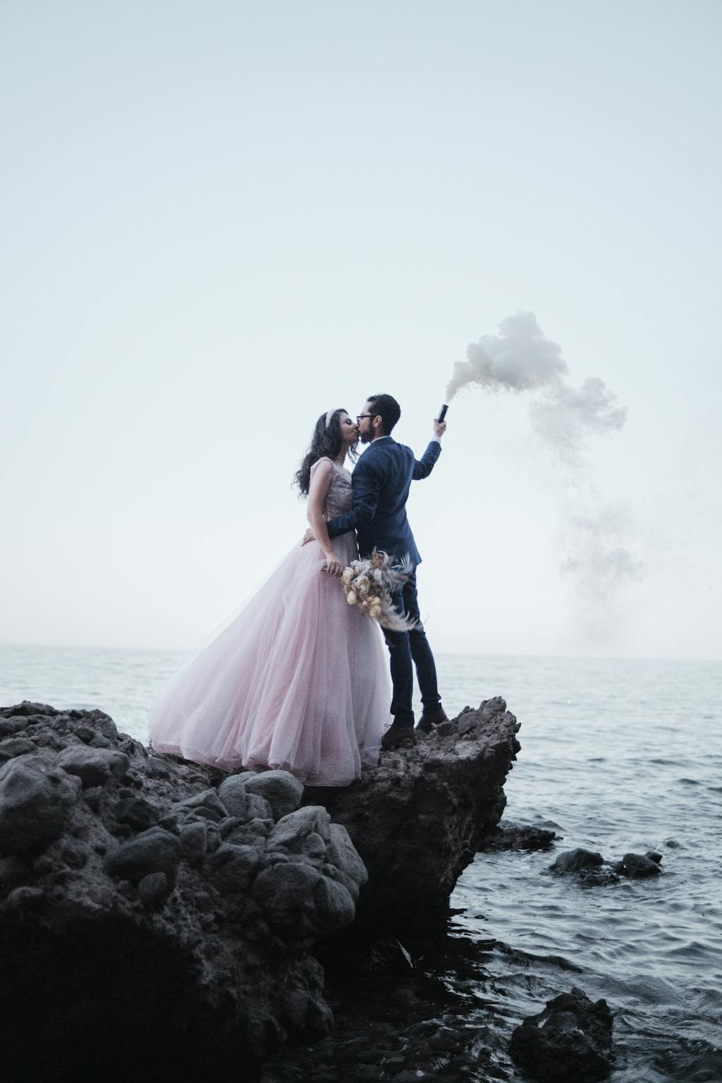 A bride and groom stand on a rocky coast, holding a smoke bomb against the ocean backdrop.