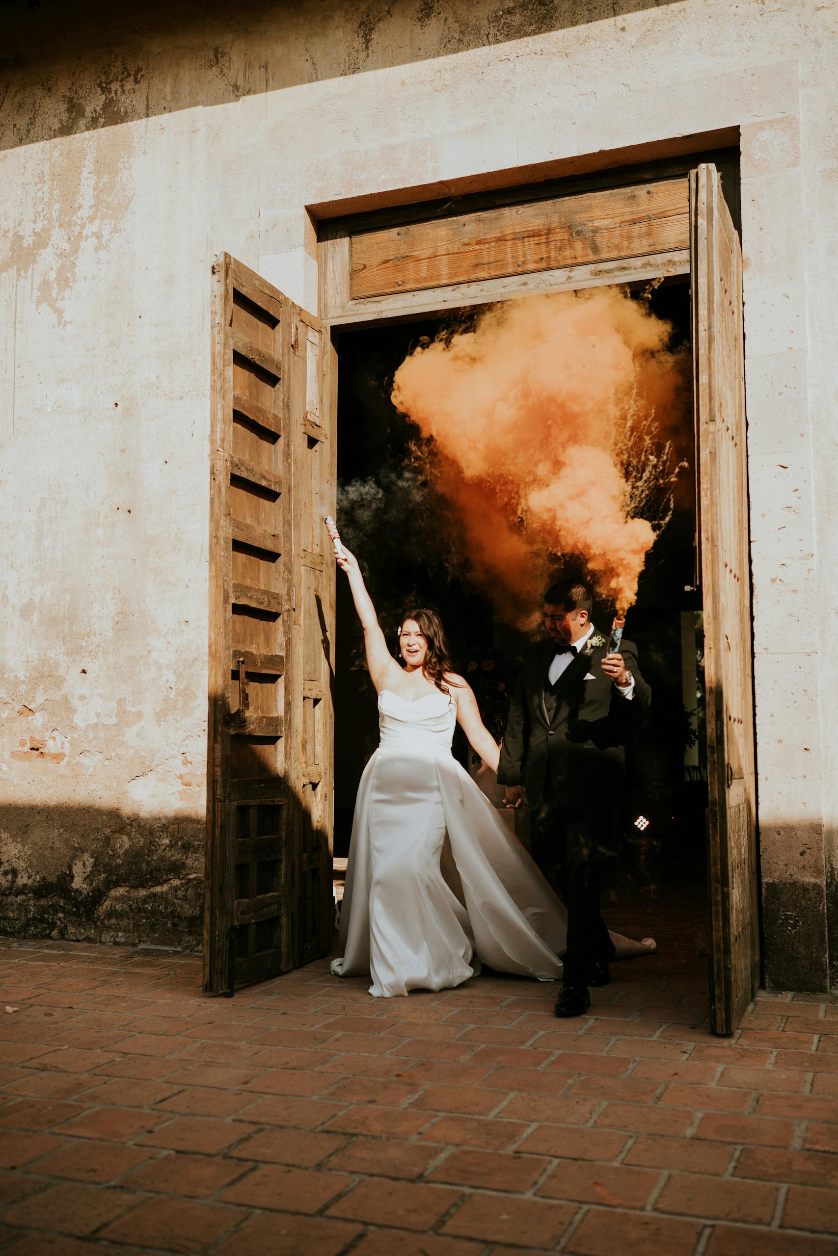 Bride and groom make a vibrant exit with colorful orange smoke at rustic wedding venue.