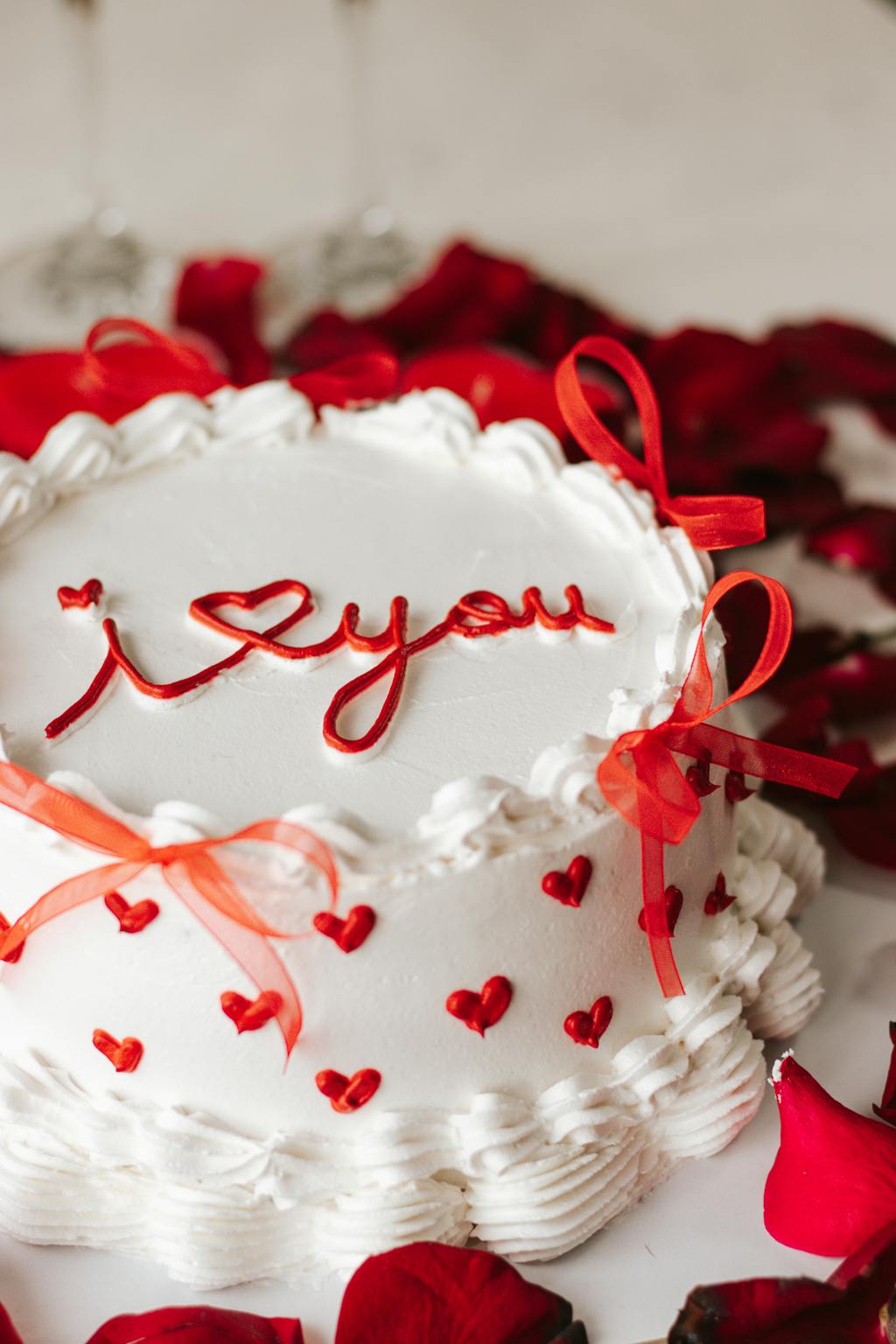 White wedding cake with I love you icing, red bows, surrounded by rose petals, ideal for romantic occasions.