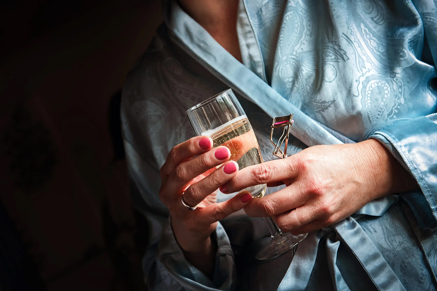 Bride getting ready moment holding champagne and a lash curler in her hand.