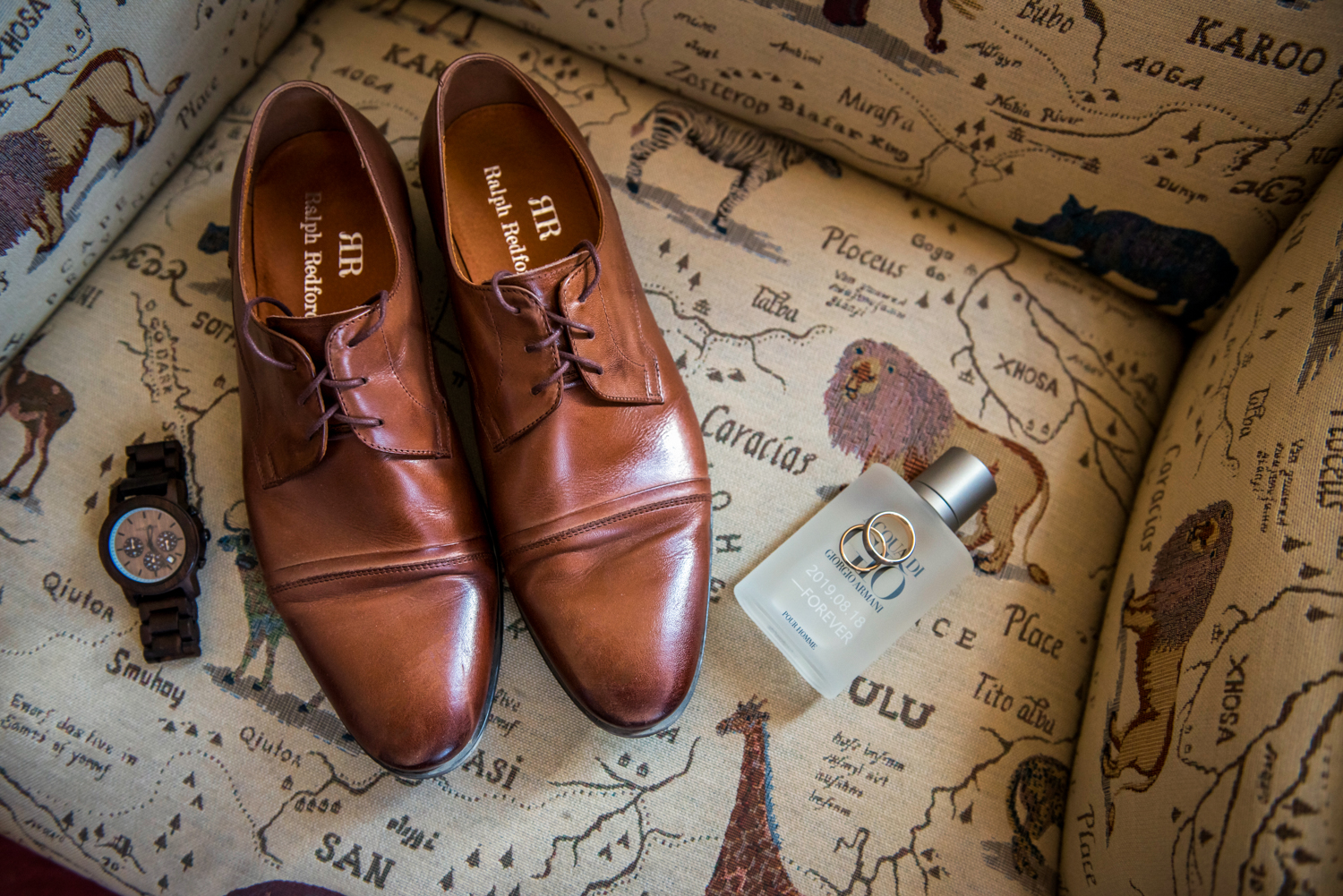 The groom is getting ready with accessories such as personalised Giorgio Armani perfume, a wooden watch, and Ralph Redford shoes on an animal pattern sofa.