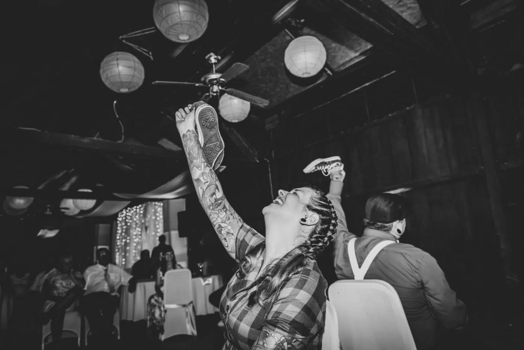 A tattooed bride laughing during a playful wedding shoe game surrounded by cheering guests.