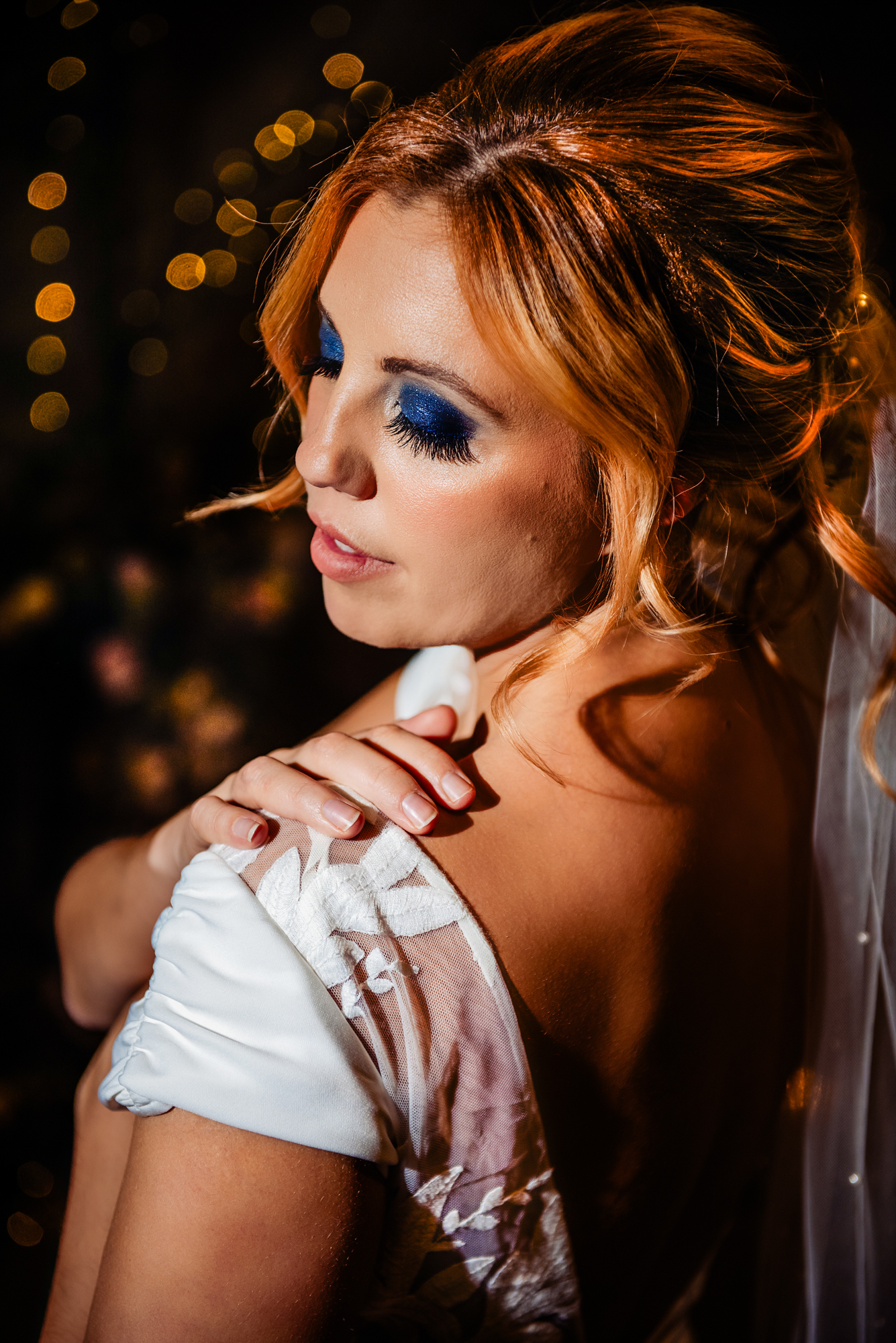 Bride resting her hand on her shoulder, showcasing bold blue eyeshadow and soft editorial lighting.