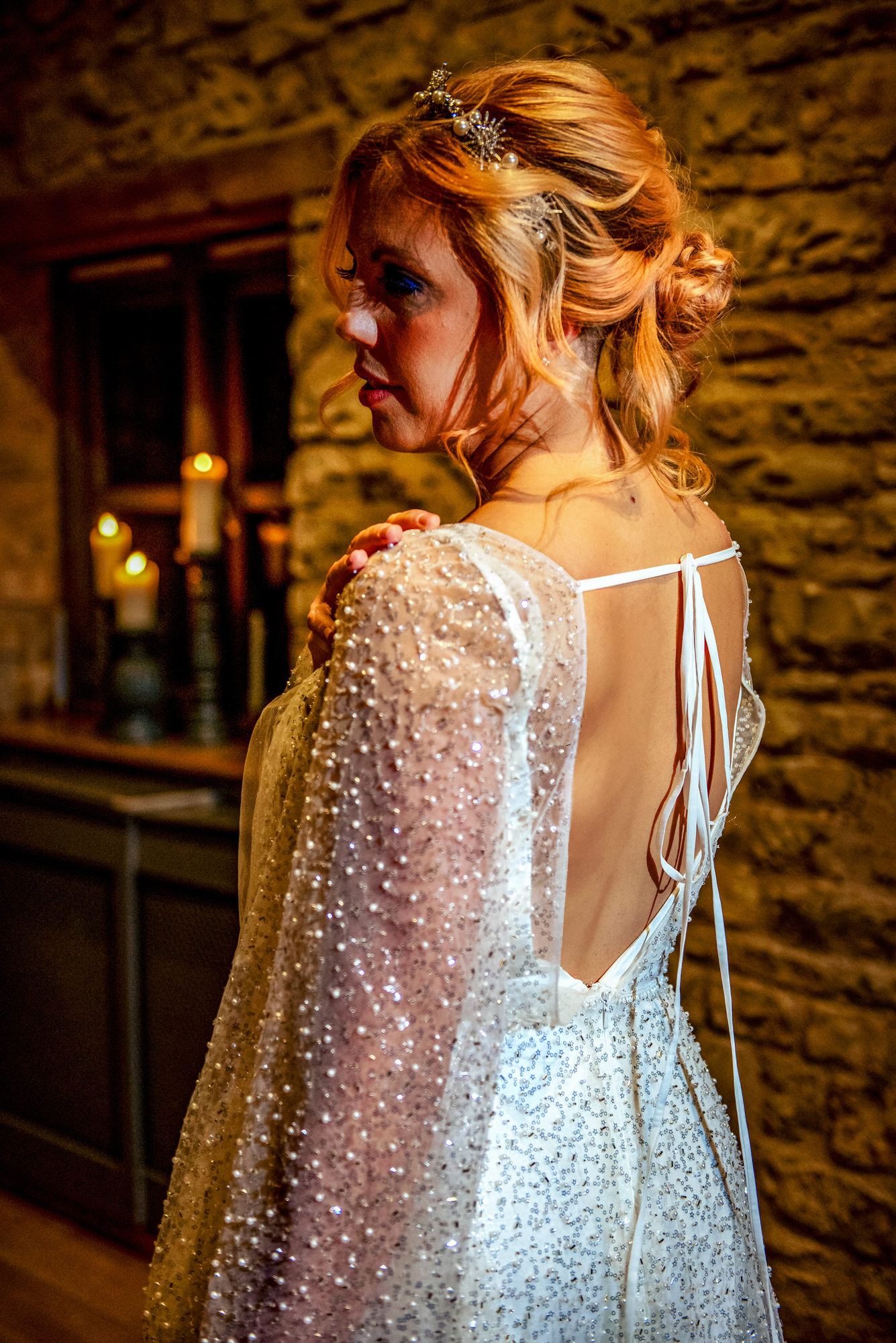 Back view of the bride’s open-back gown as candlelight reflects on her pearl-detailed sleeves.