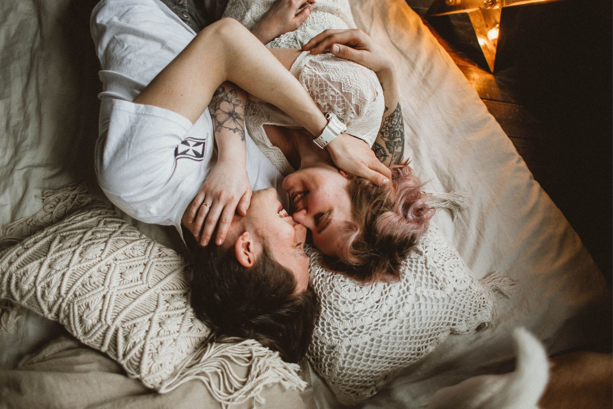A couple is laughing together during a cosy indoor couple photoshoot with soft blankets and warm light.