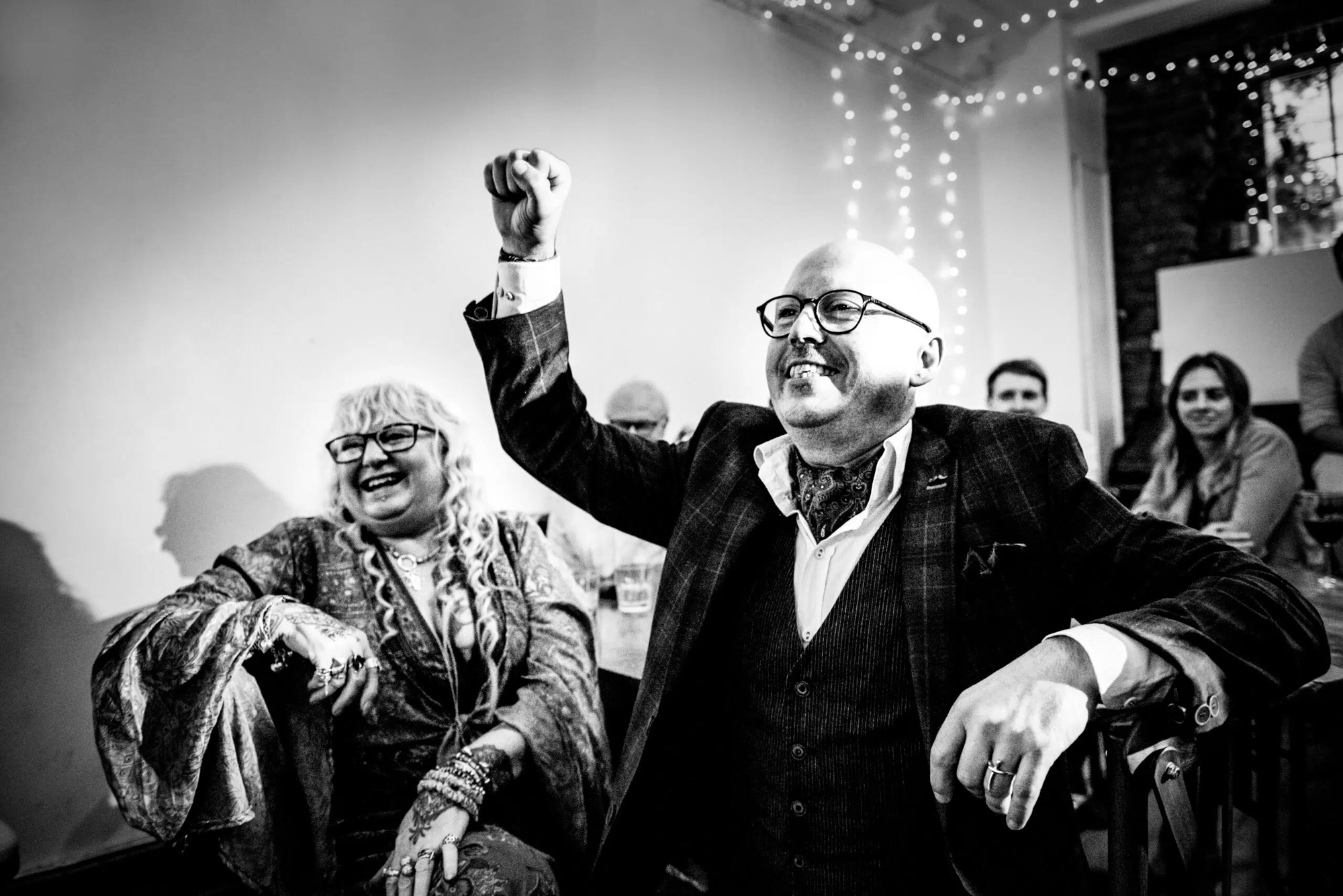 Guest raising his arm laughing during emotional wedding breakfast speeches with family nearby.