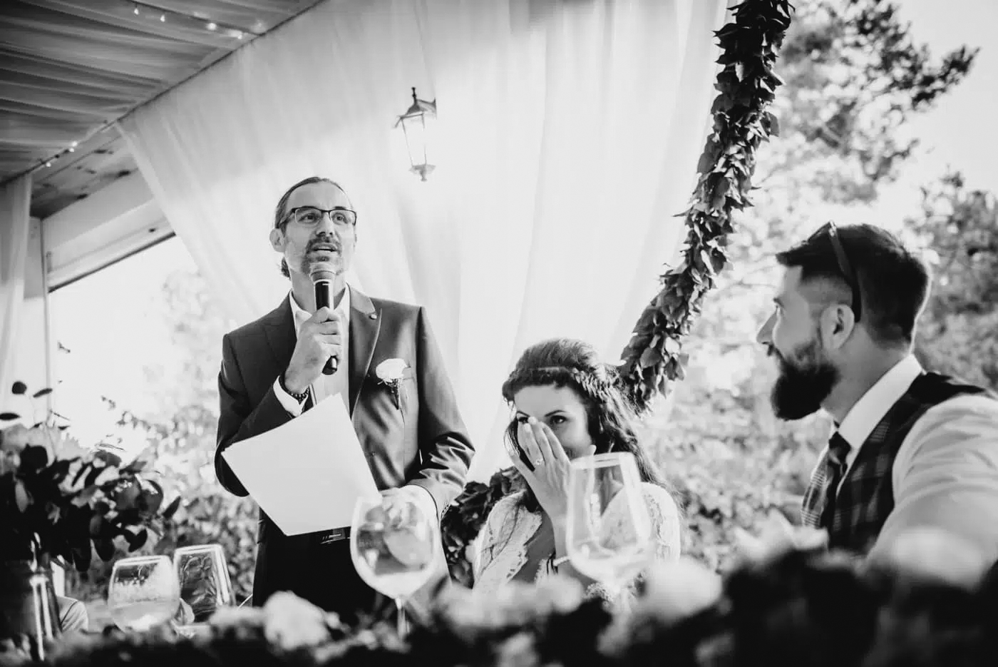 Best man delivering speech during wedding breakfast while couple react with emotion at top table.