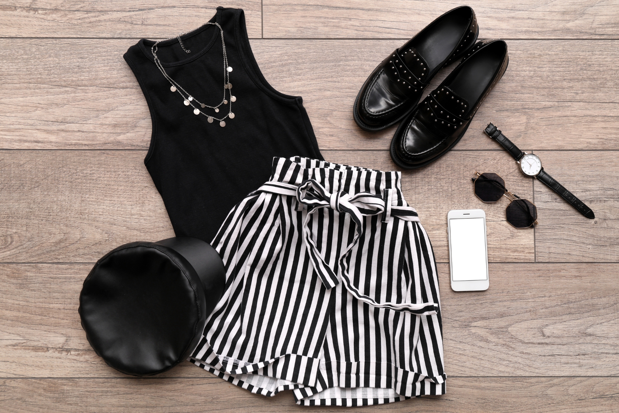 Flat lay outfit with black sleeveless top, striped skirt, shoes and accessories on wooden floor.