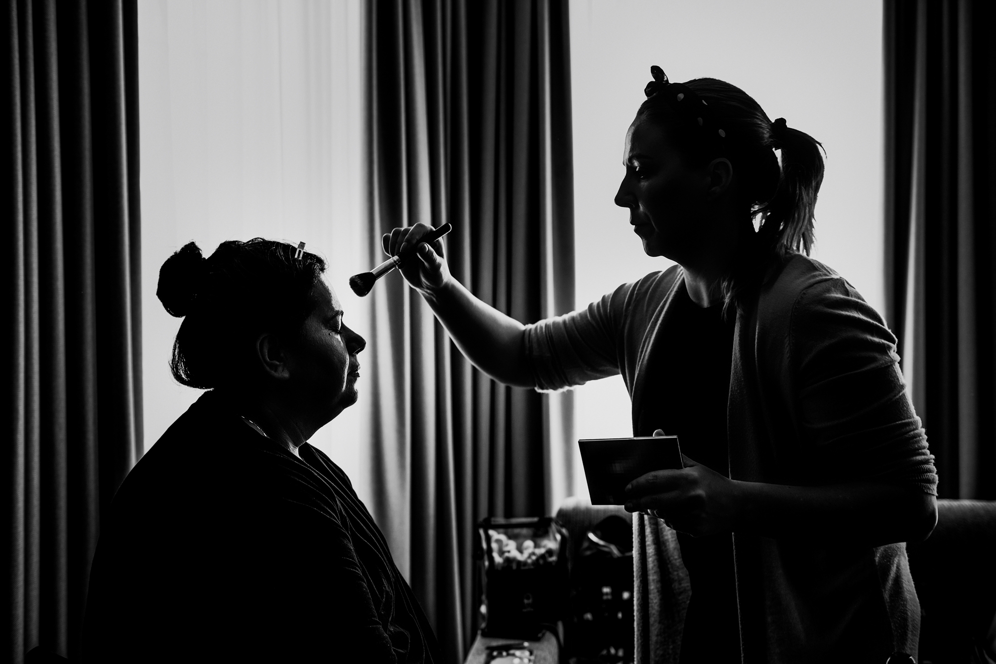 Bride's mum getting makeup done by window light at Arnos Manor Hotel Bristol wedding morning preparation.