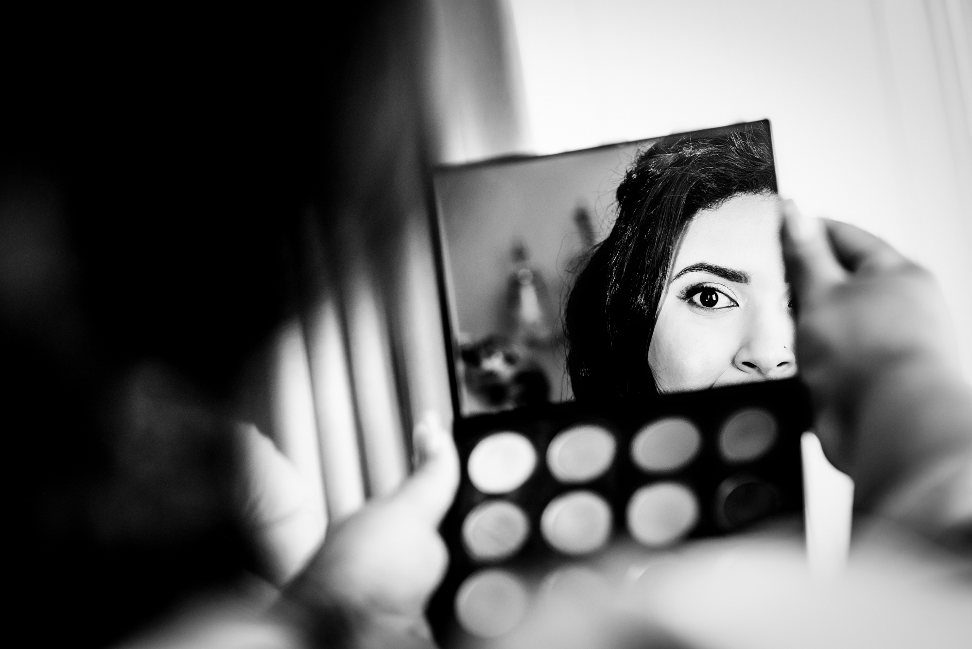Creative reflection shot of bride applying makeup using mirror.