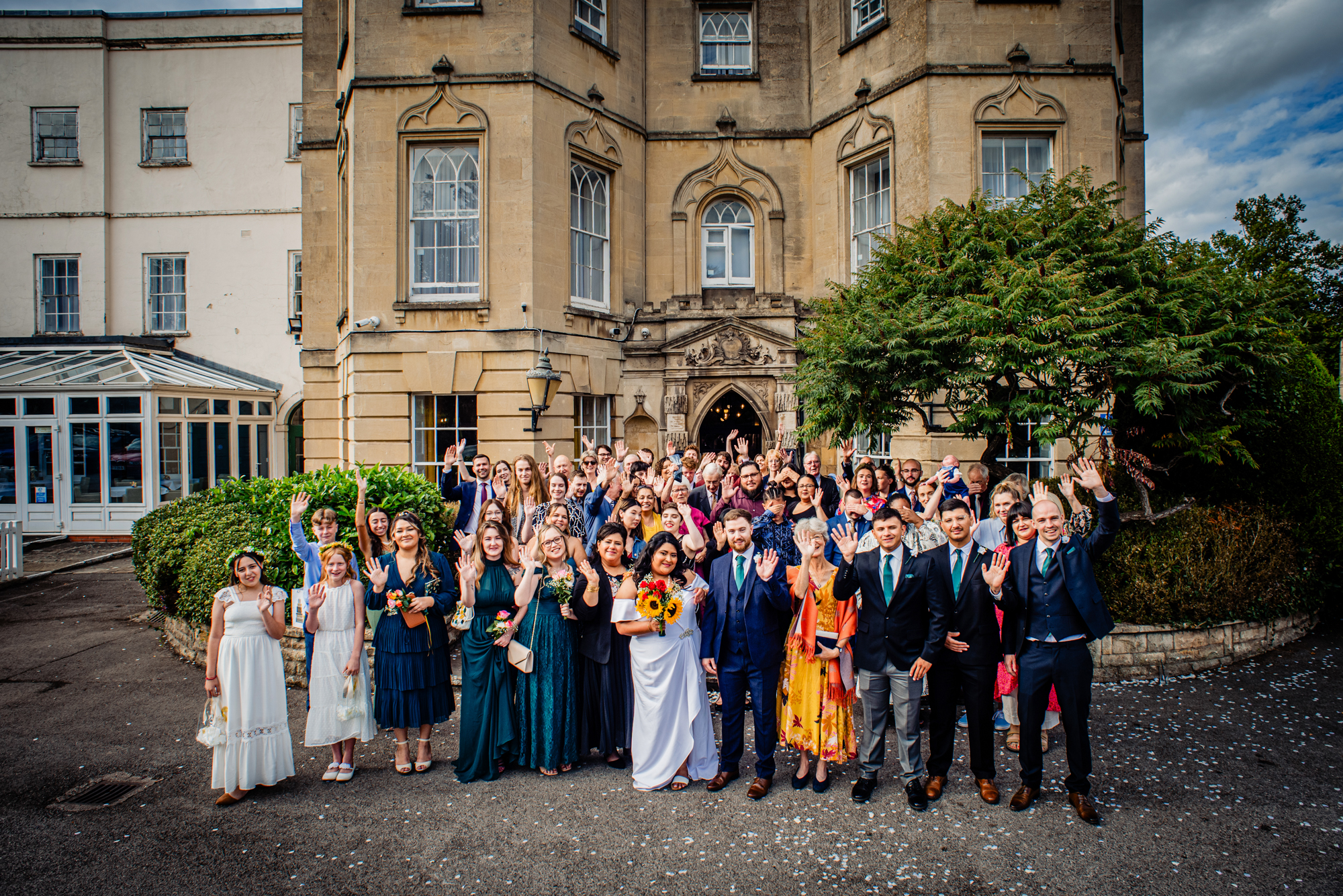 Large group photo of wedding guests waving outside Arnos Manor Hotel in Bristol.
