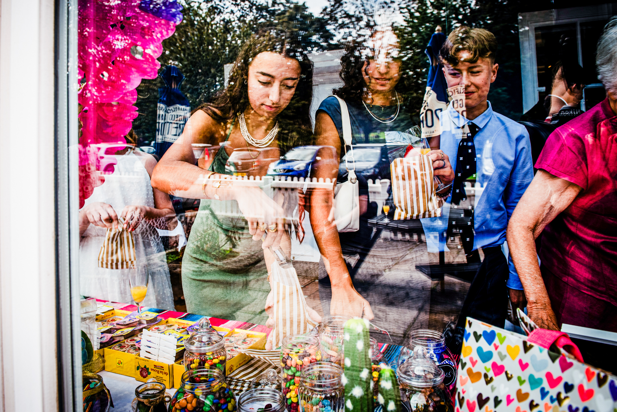 Guests choosing sweets at colourful wedding candy table through window reflection.