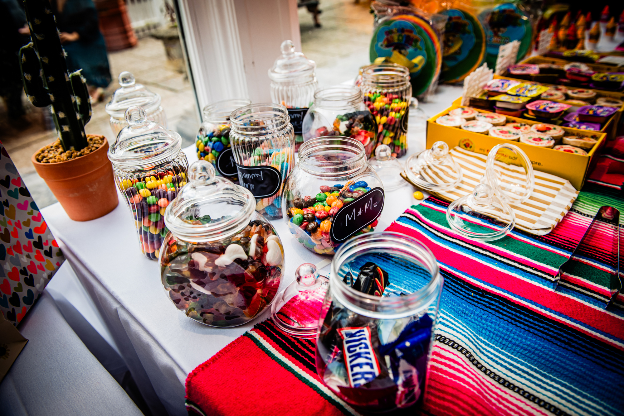 Glass jars filled with sweets arranged on vibrant wedding dessert table with M&Ms.