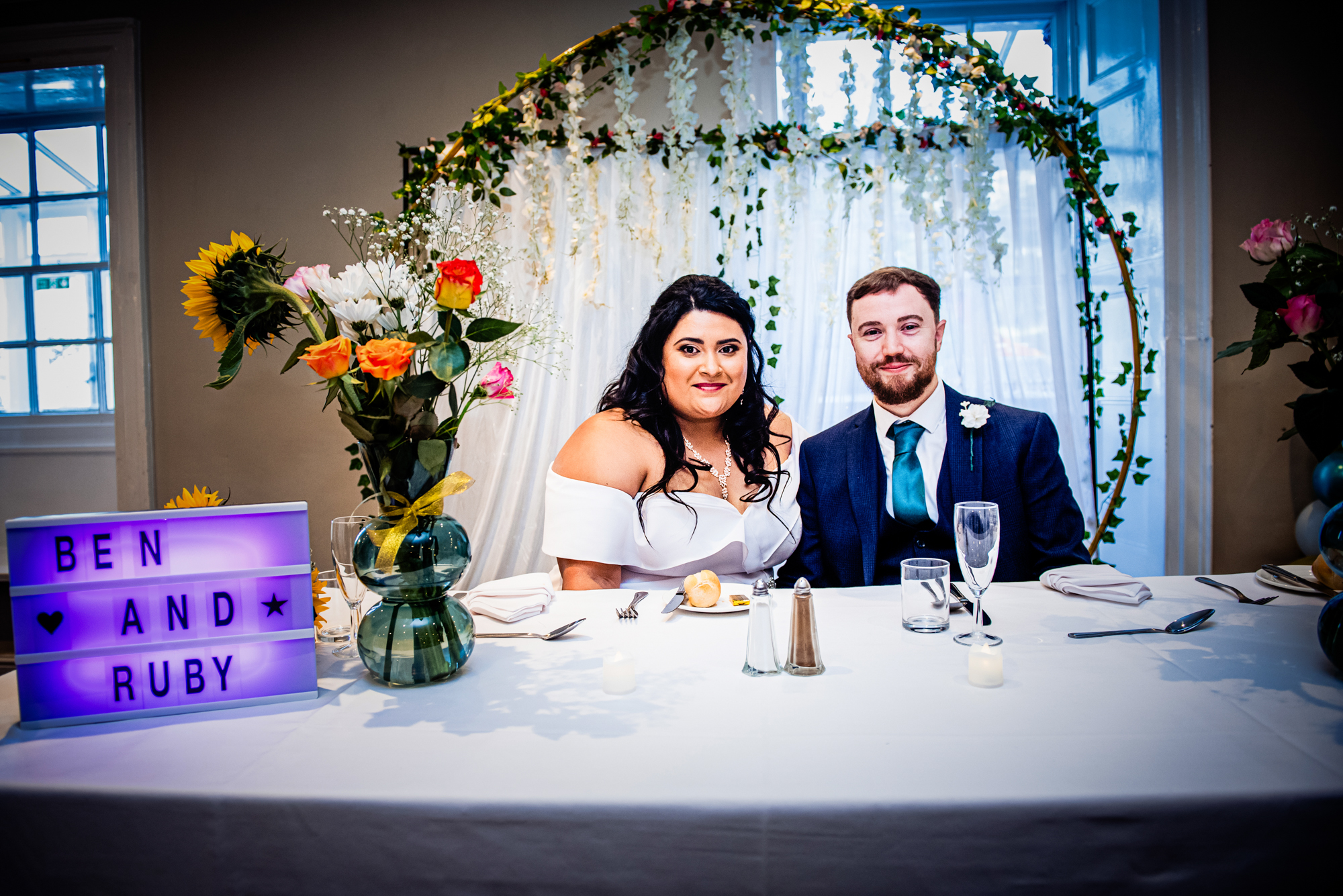 Couple seated at decorated top table with floral arch backdrop.