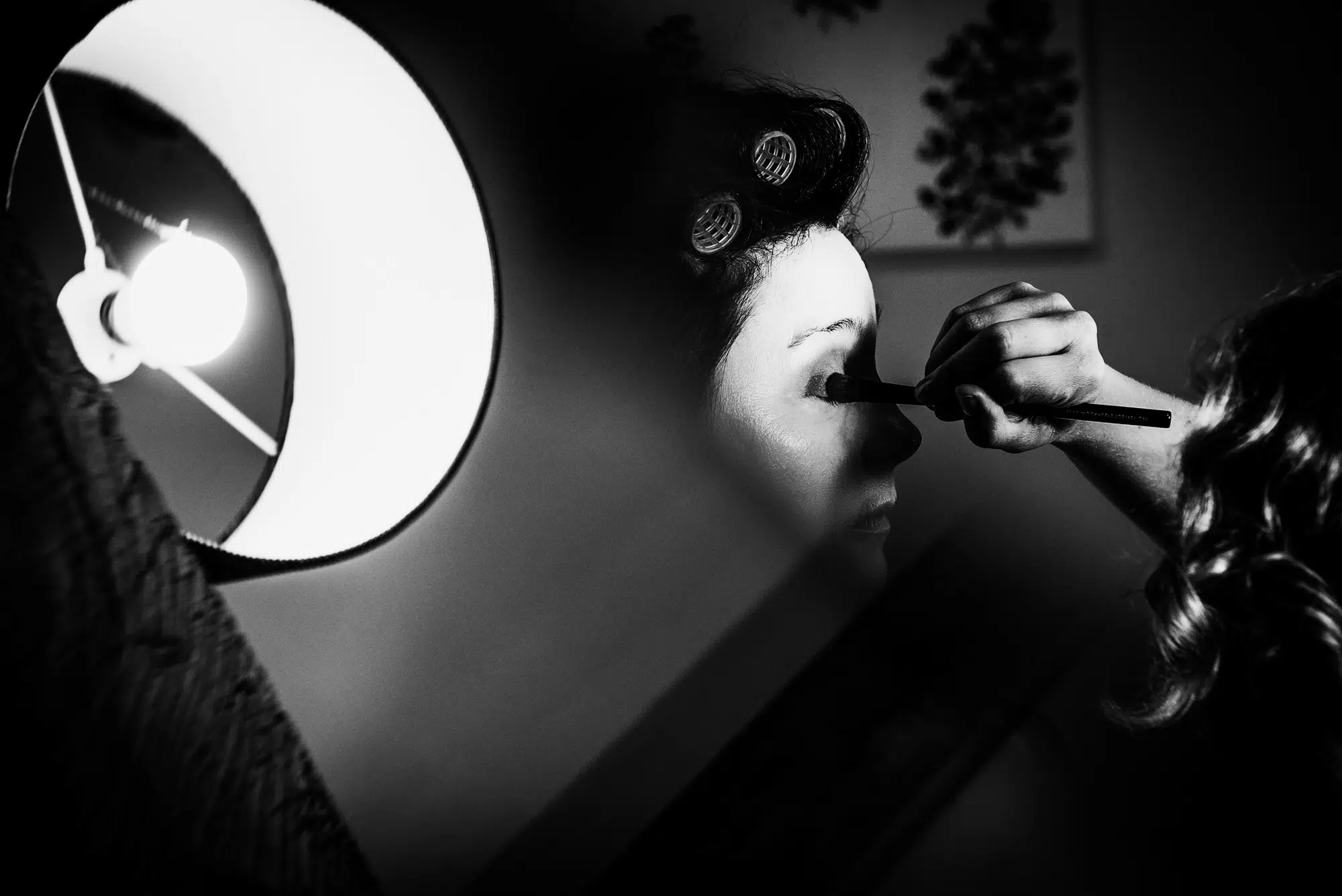 Bride having makeup applied in dramatic window light during wedding morning preparations.