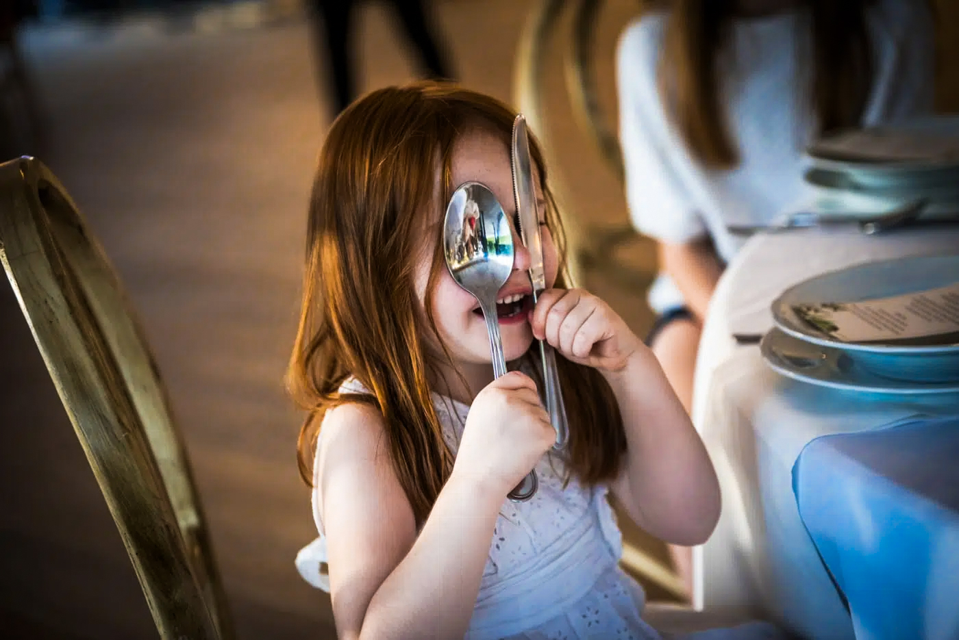 Young flower girl playfully holding spoons at wedding breakfast table.