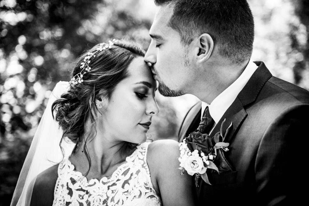 Groom kissing bride’s forehead, intimate black and white wedding moment outdoors.