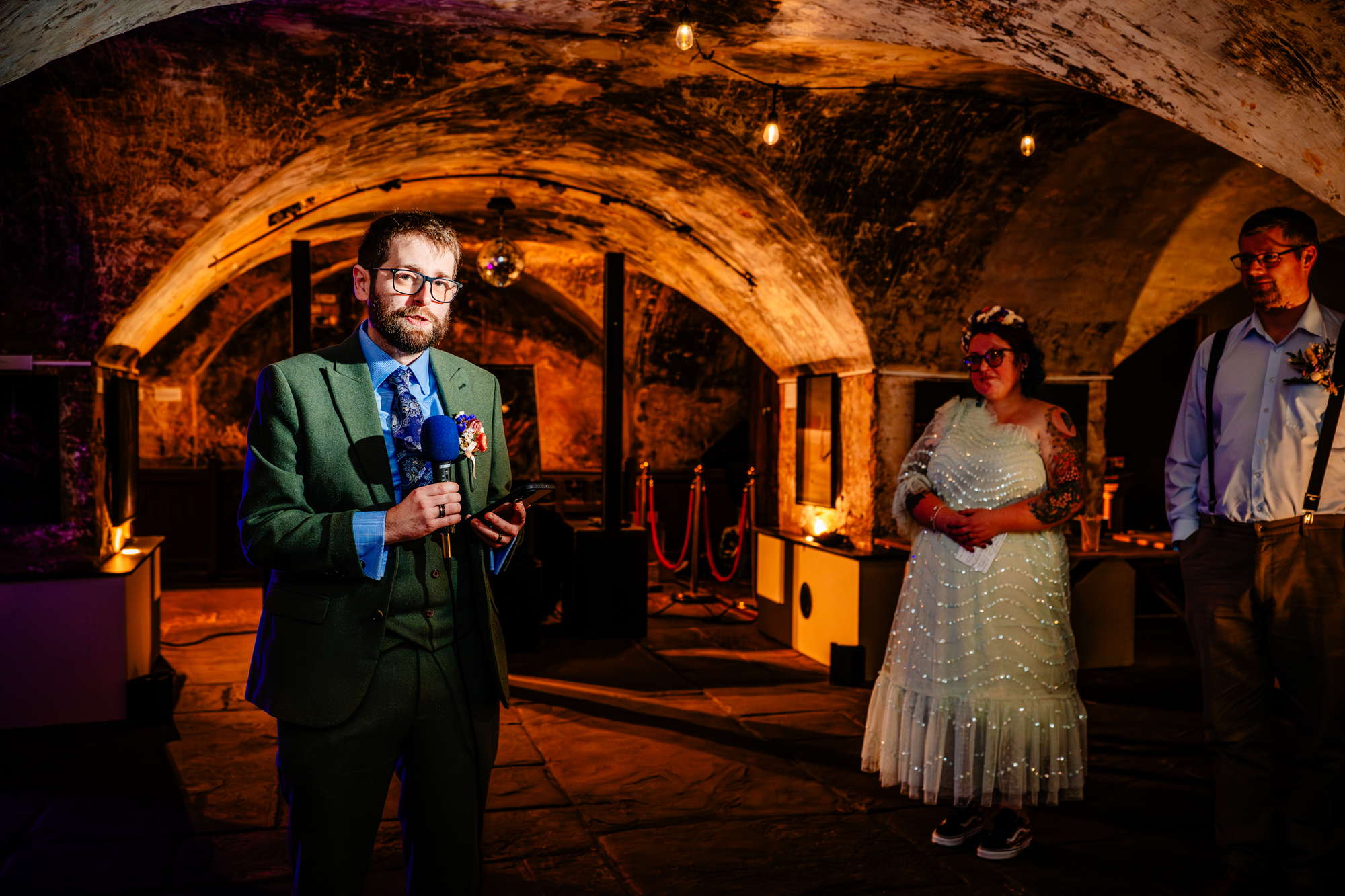Groom in a suit gives a speech with a microphone while others watch in a vaulted, dimly lit room.
