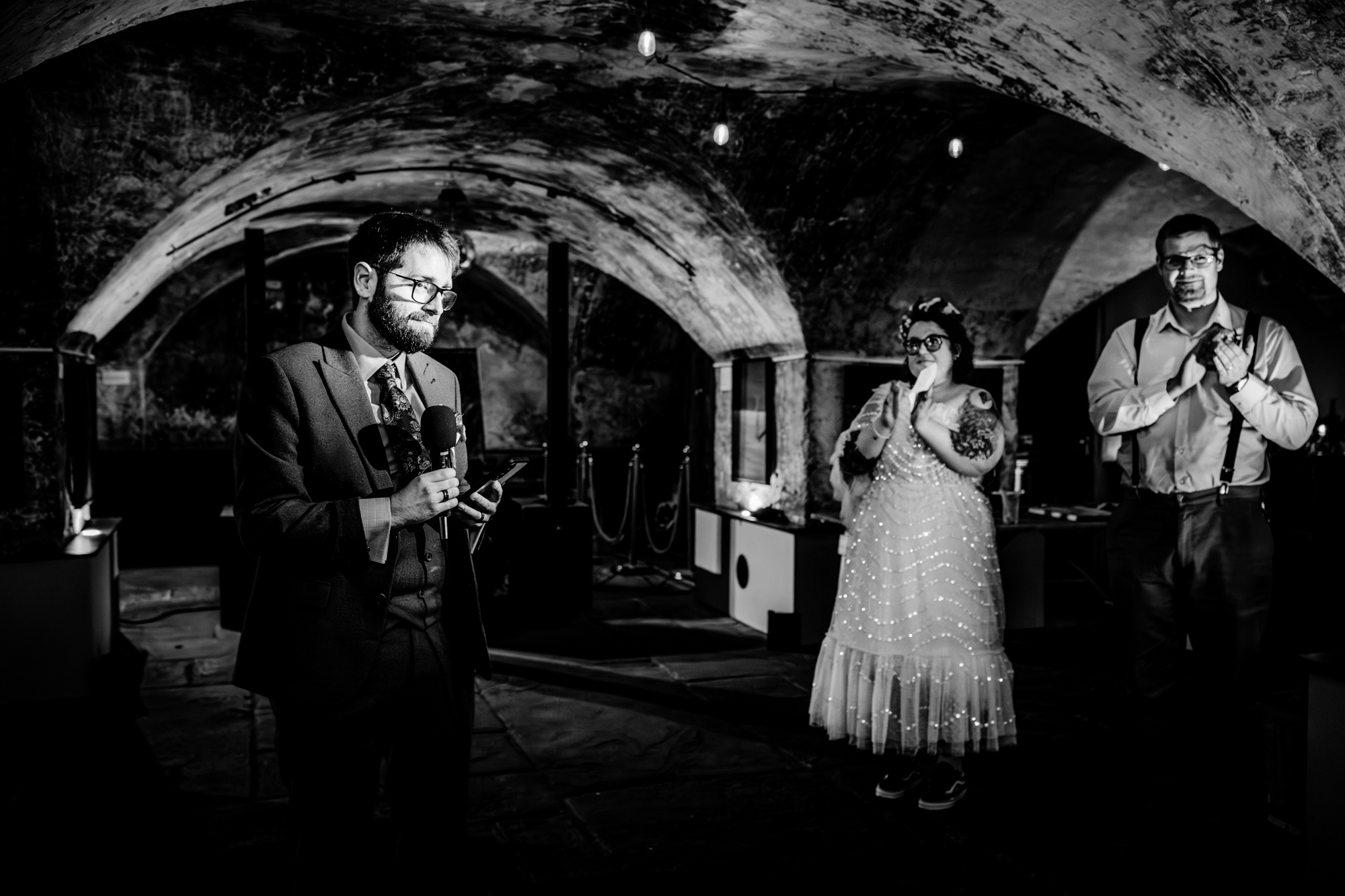 Audience of formally dressed guests watches attentively, some holding drinks, in a warmly lit space.