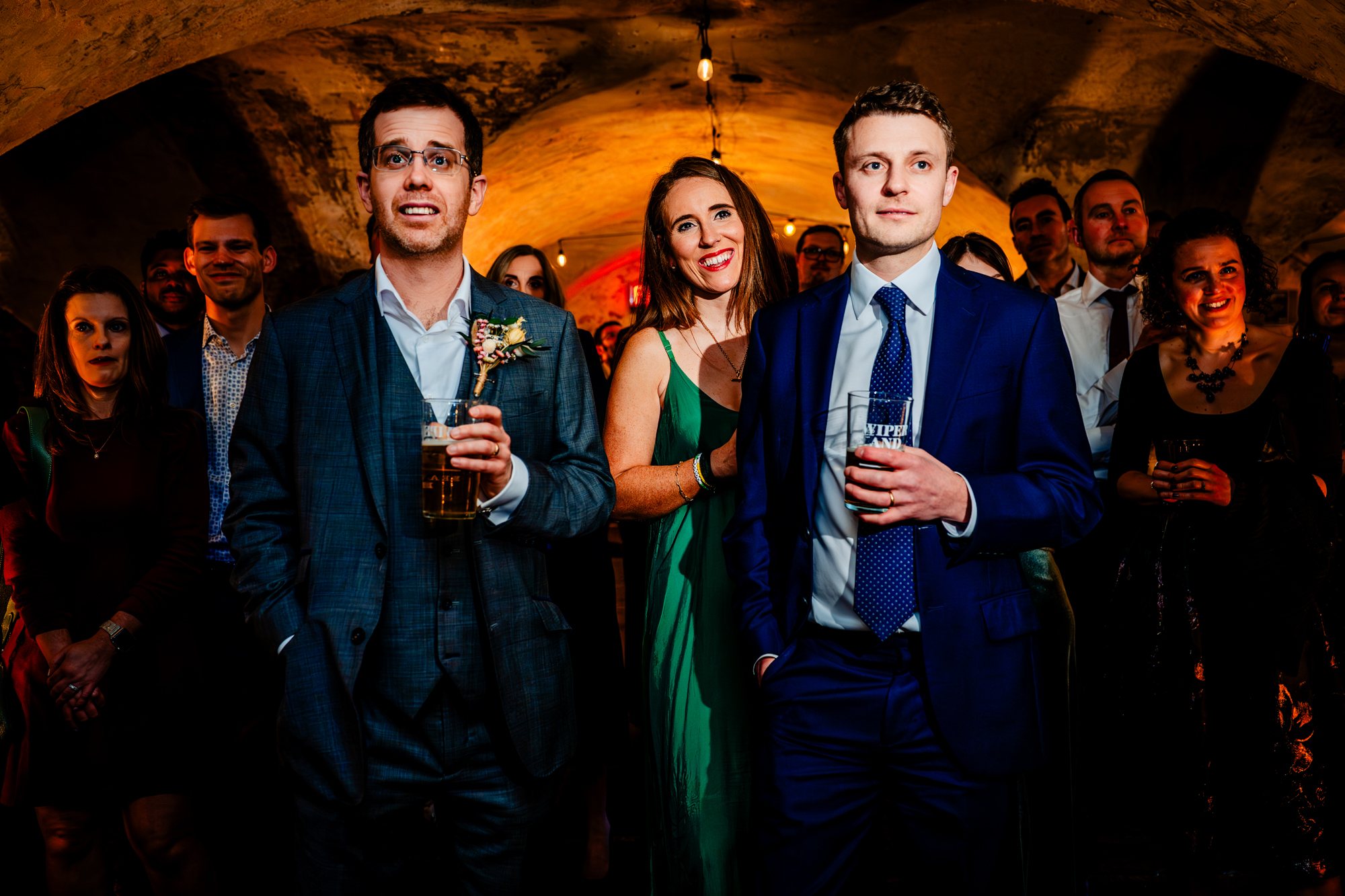 Wedding guests in formal attire smile at each other during a wedding speech, standing under arched ceilings.
