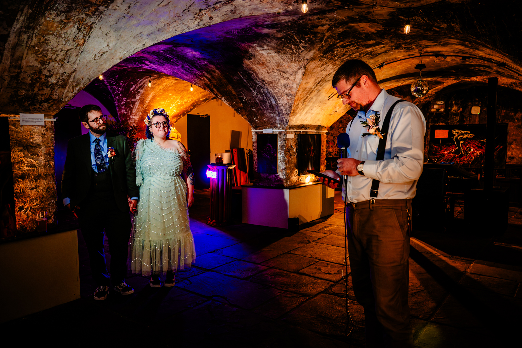Close-up of a person holding a microphone and phone while speaking, with two others watching behind.