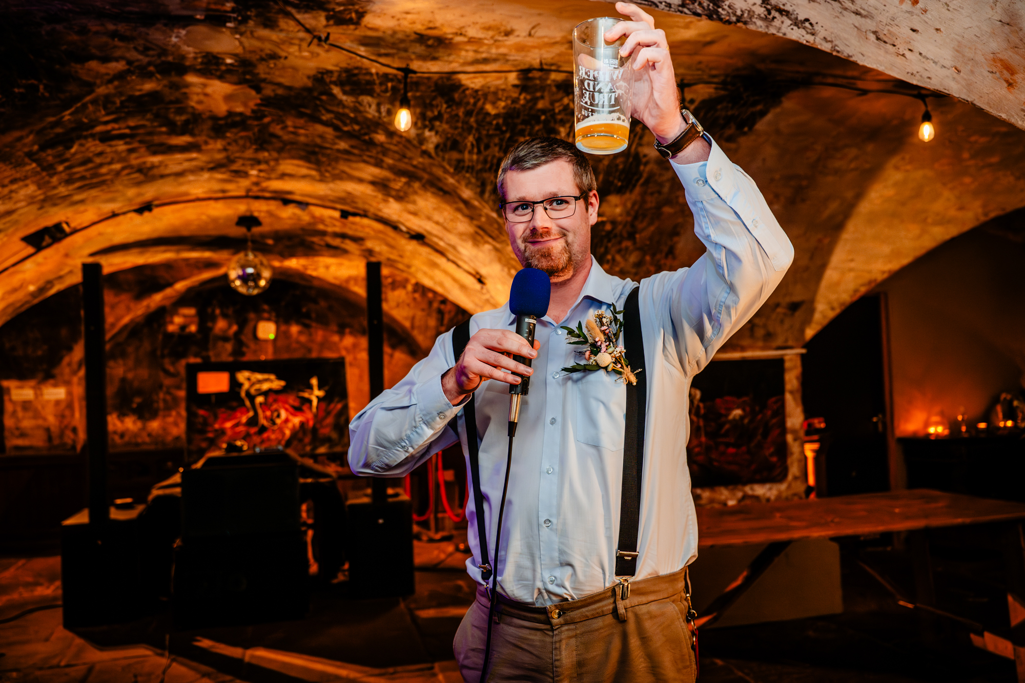 Person raises a glass mid-toast, smiling and addressing the room with a microphone.