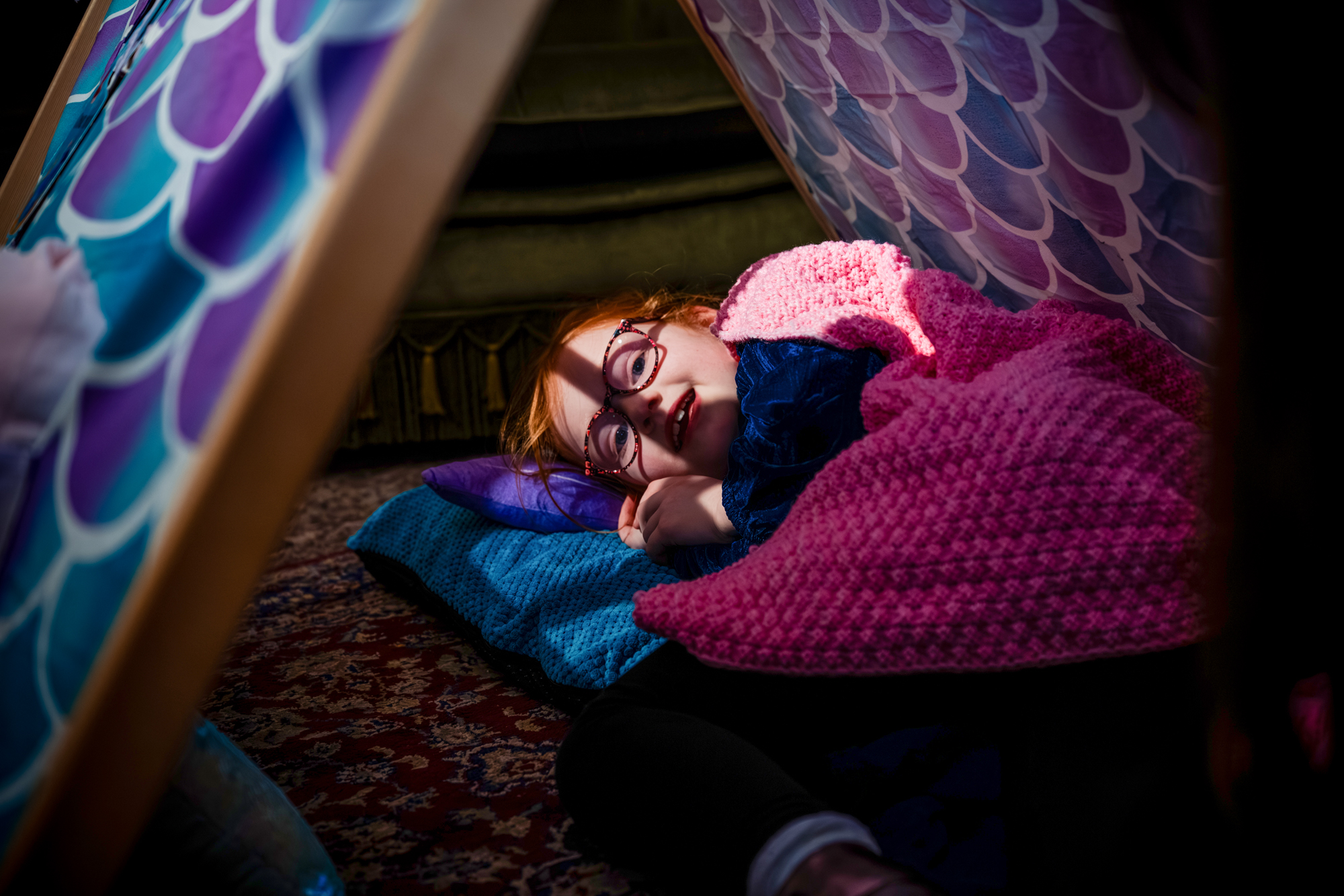 Child with glasses lies under a small blanket fort, smiling while resting on cushions at the Mount Without in Bristol