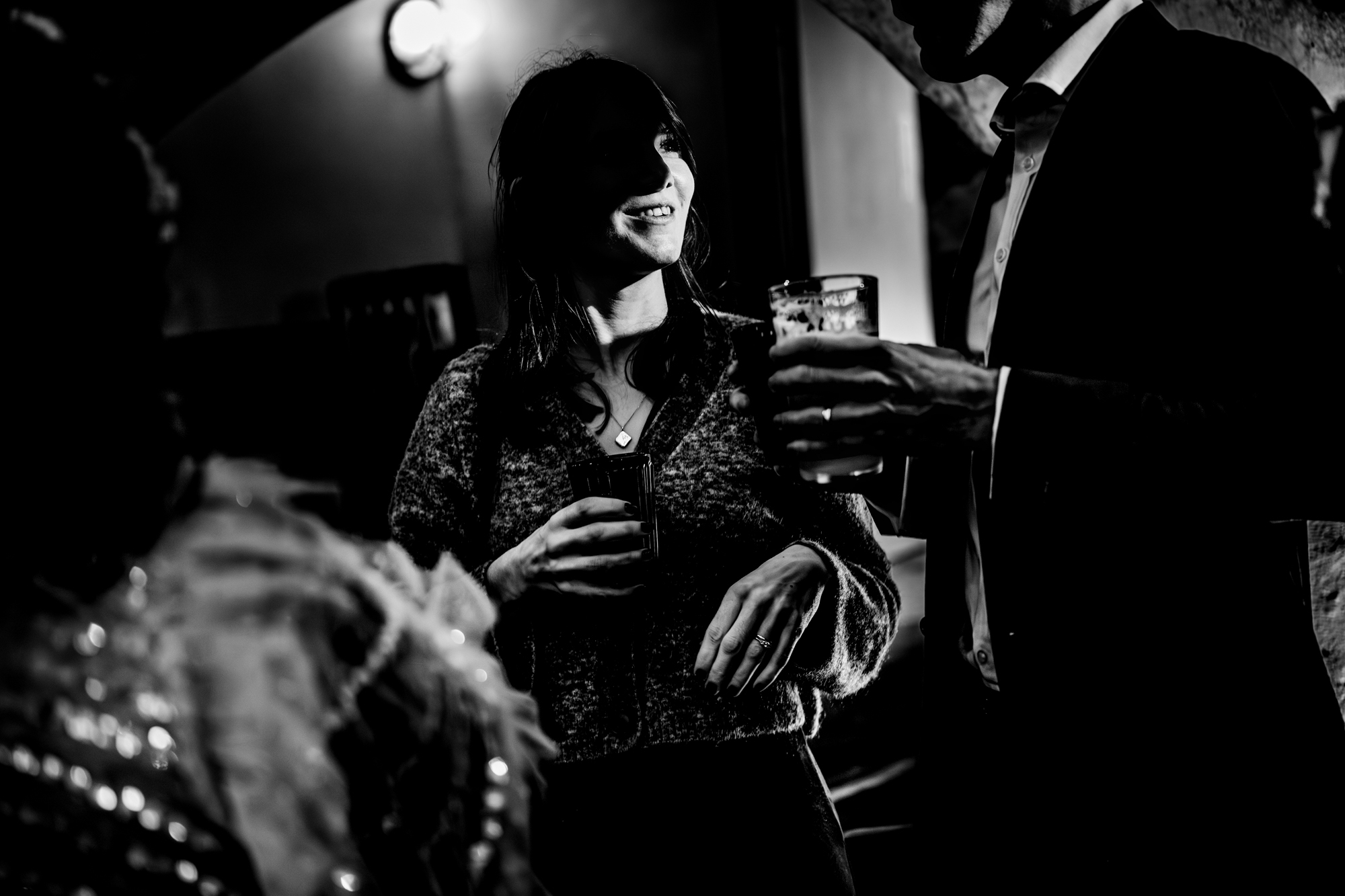 Two wedding guests in dim lighting hold drinks and talk closely, partially silhouetted.
