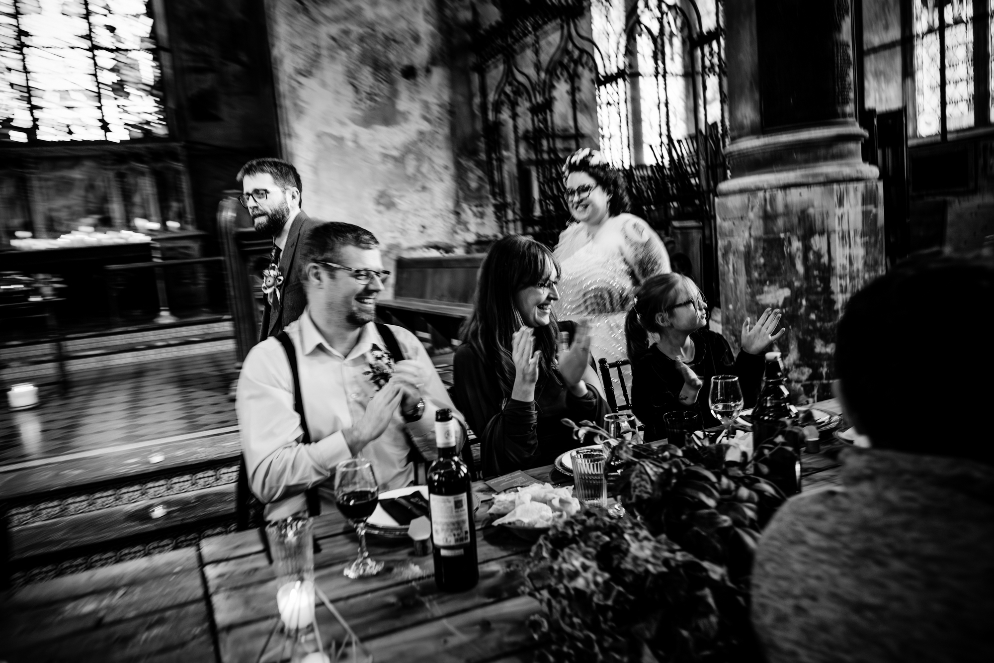 Wedding guests seated at a decorated table clap and smile as the couple passes behind them.
